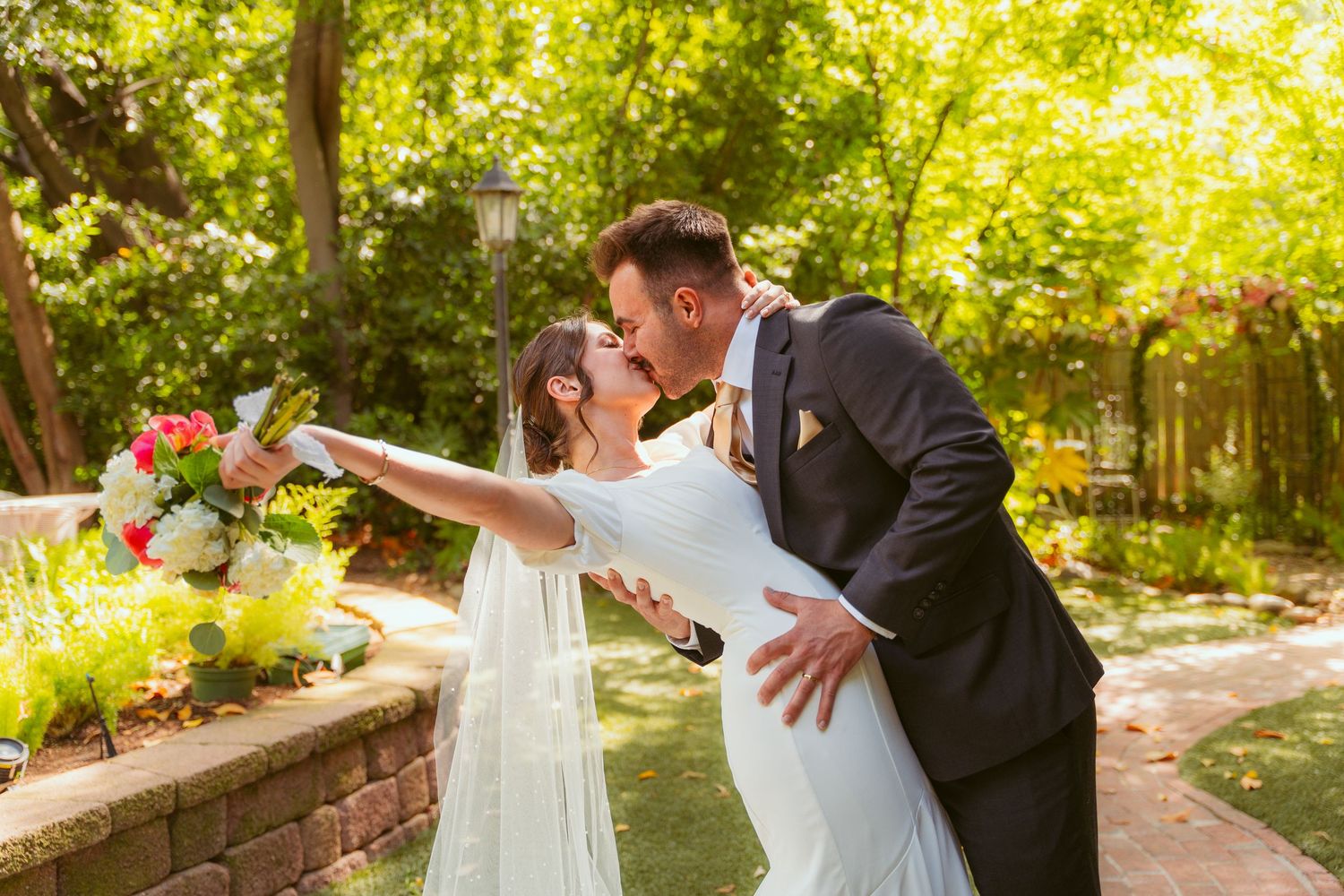 A playful wedding dip pose in a garden setting with natural sunlight filtering through trees.