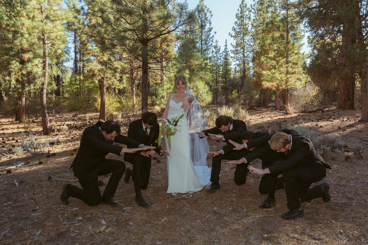 Wedding party members pose casually on ground in wooded outdoor setting.