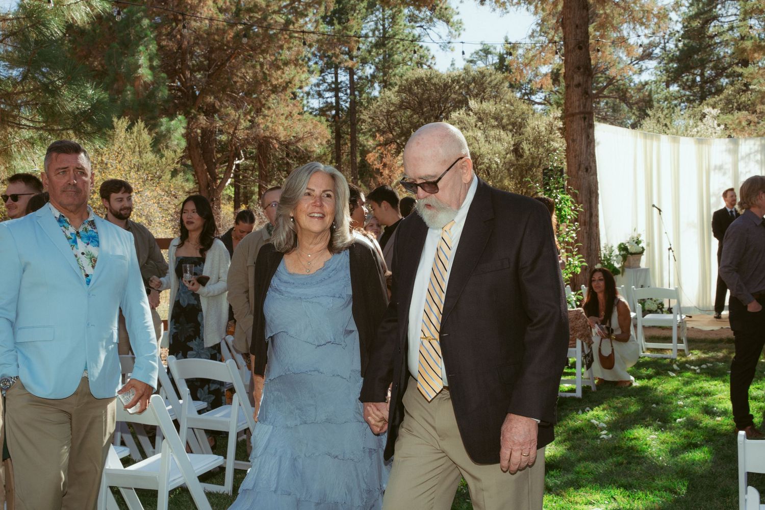 Wedding party members in formal attire gather outdoors under pine trees for photos.