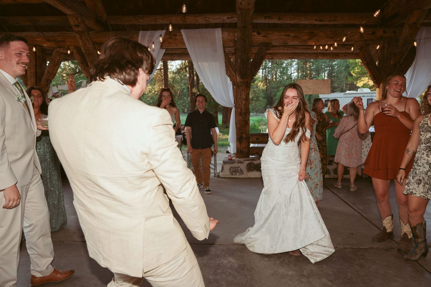 Wedding party dances in a line at an outdoor reception venue with string lights overhead.