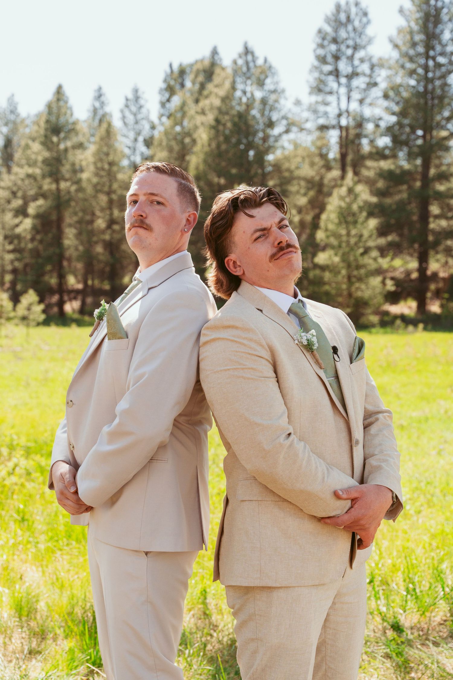 Groomsmen in cream colored suits strike humorous poses in a sunny forest clearing.