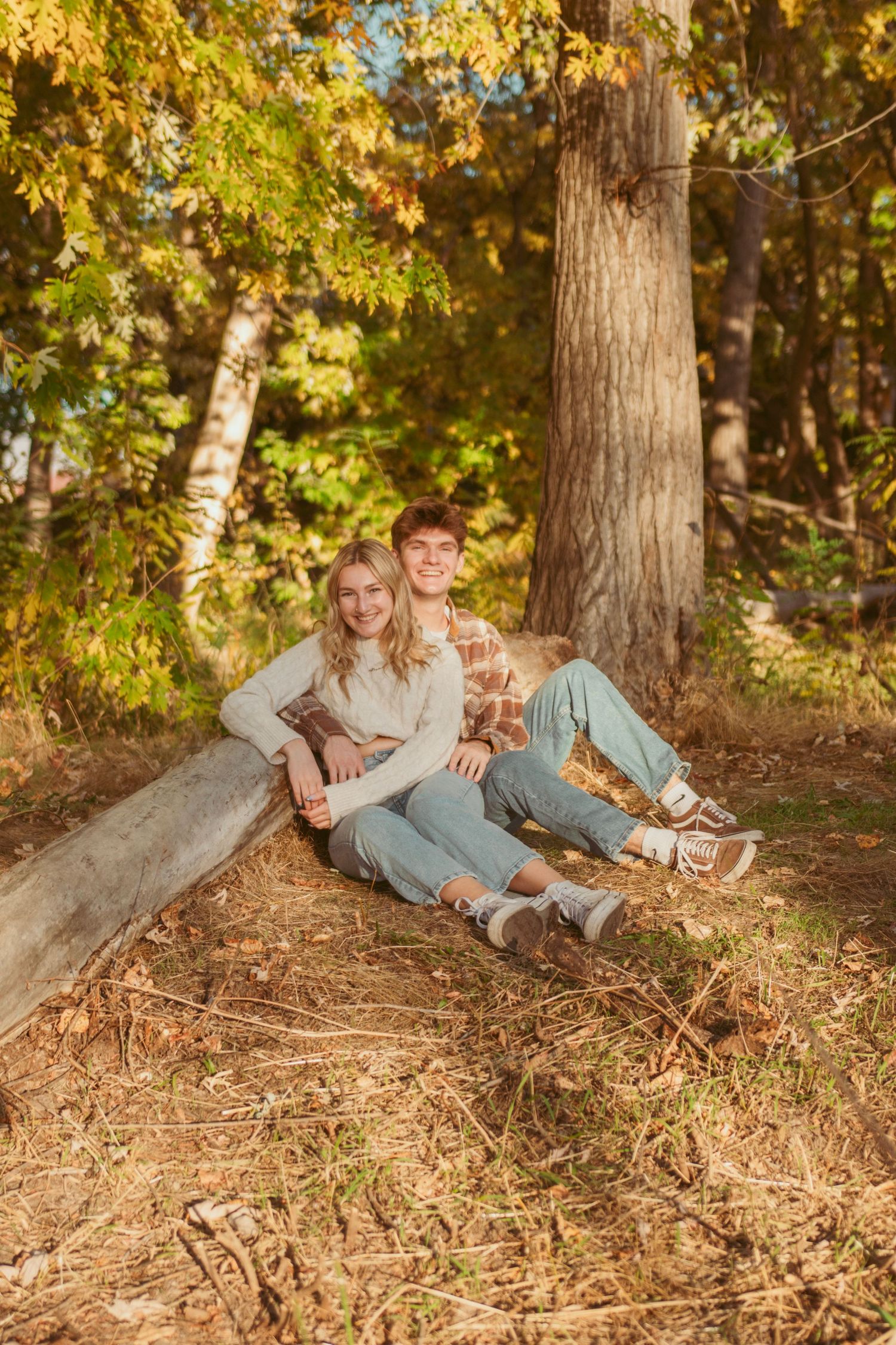 Couple relaxes on fallen leaves in autumn forest during golden hour photoshoot.