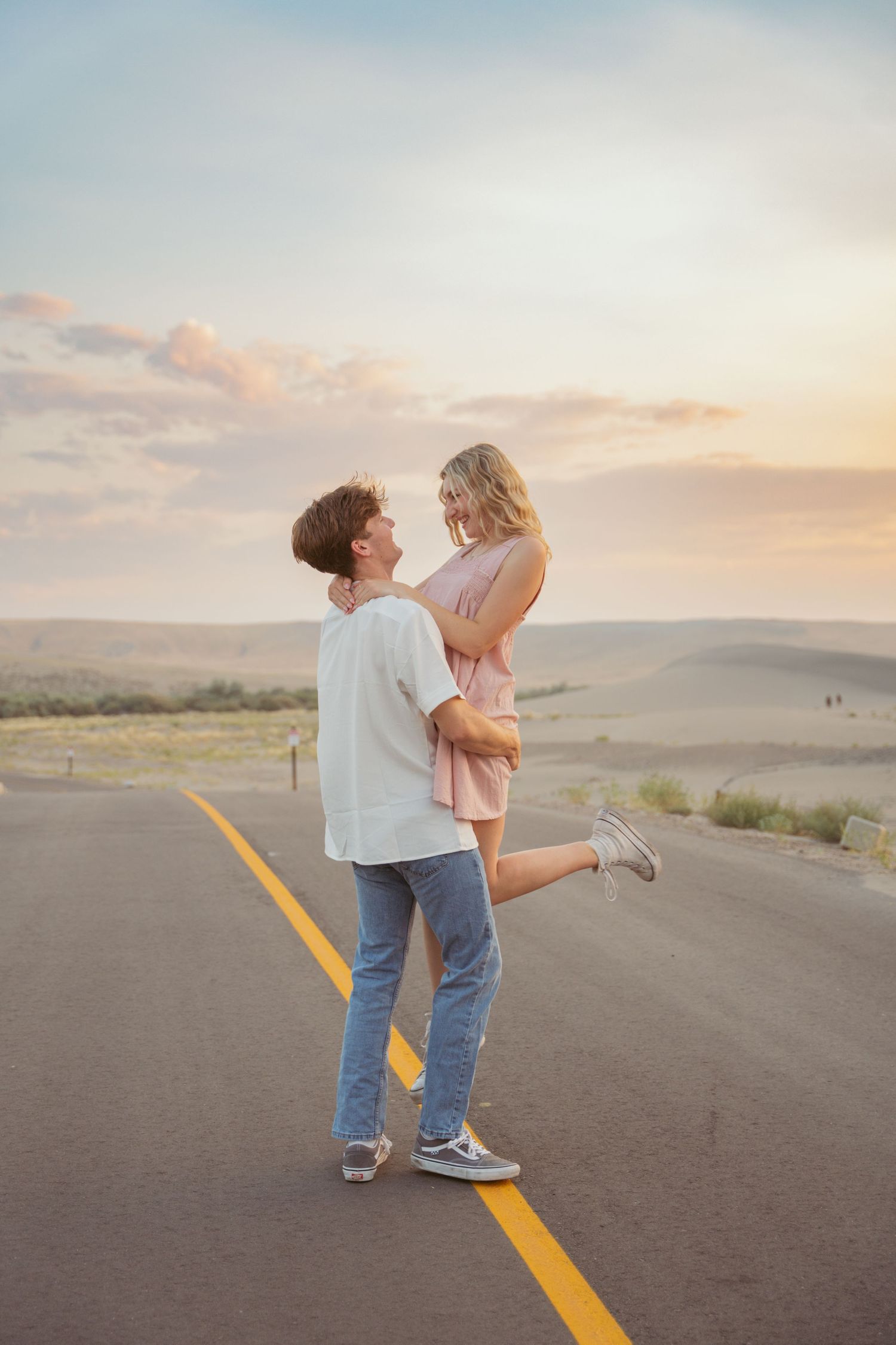 A romantic couple embraces on an empty road during sunset with rolling hills in the background.