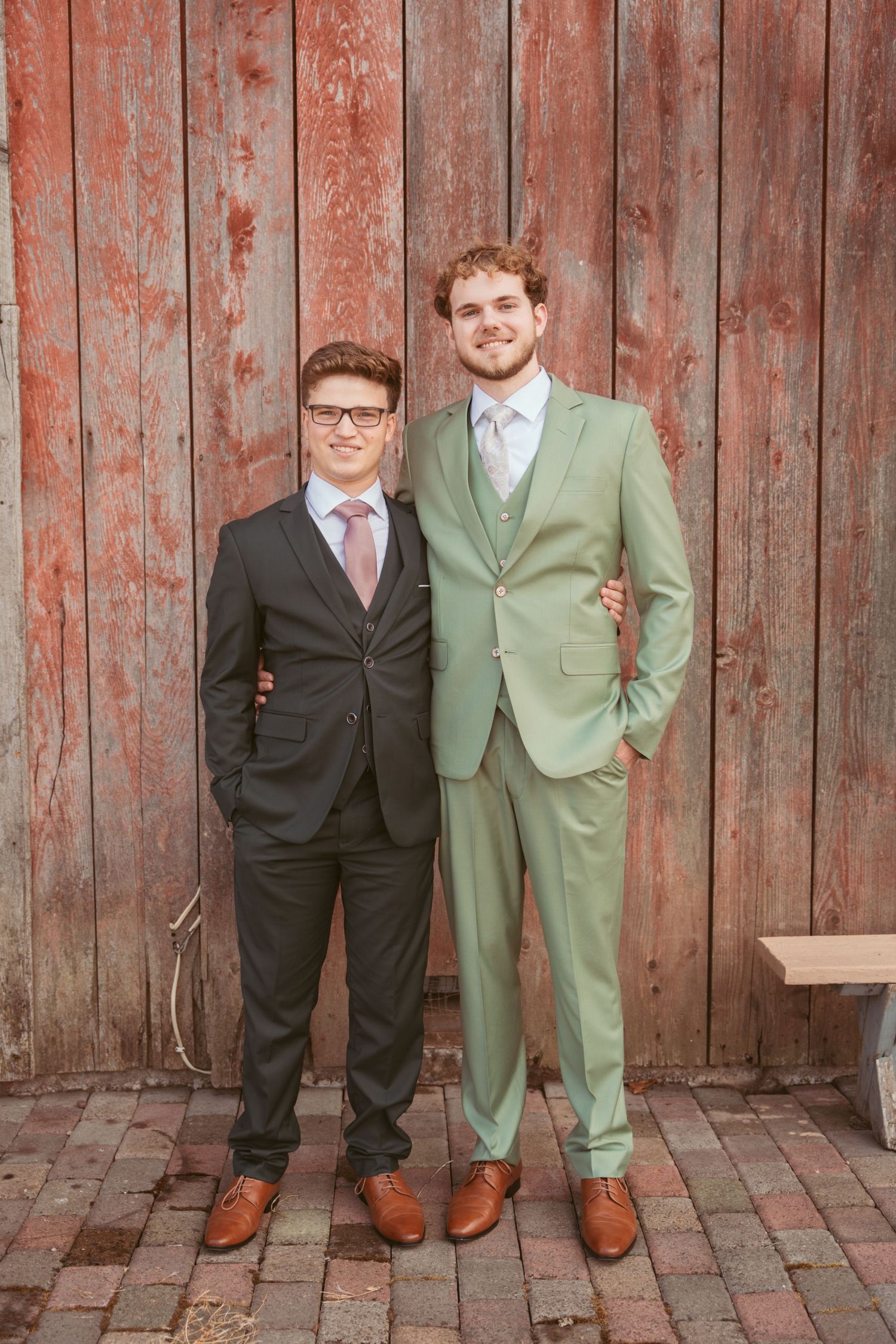 Wedding party members pose casually against a rustic red barn wall wearing black and sage green suits.