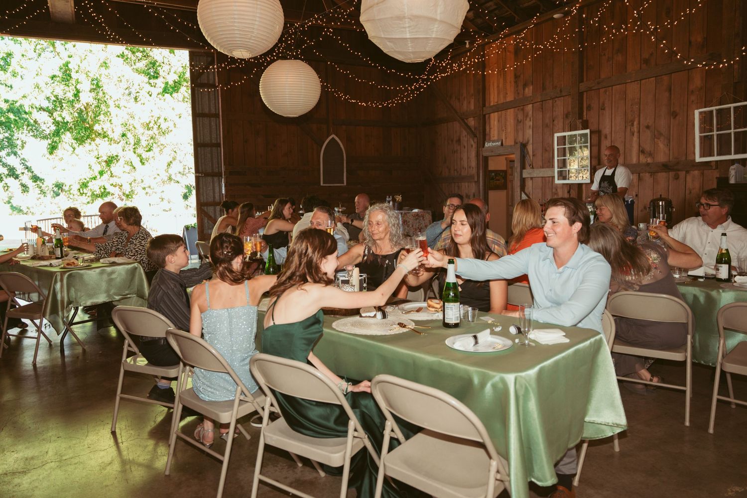 Wedding reception guests seated at round tables with green tablecloths under paper lantern decorations.