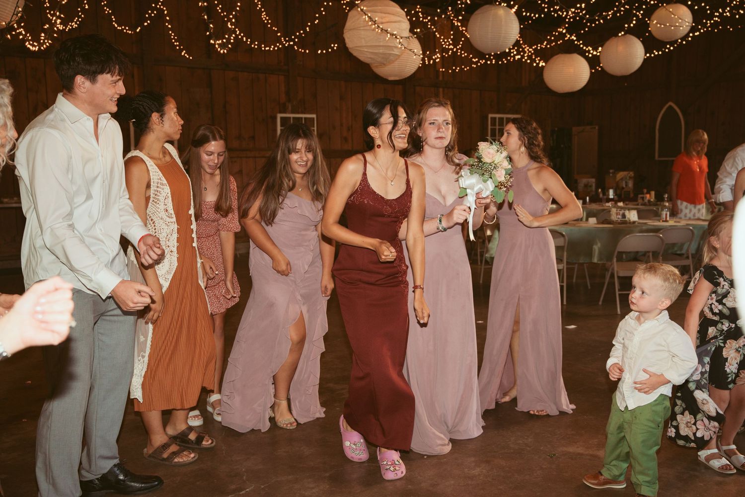 Wedding party members dancing and celebrating under warm lighting in rustic barn setting.