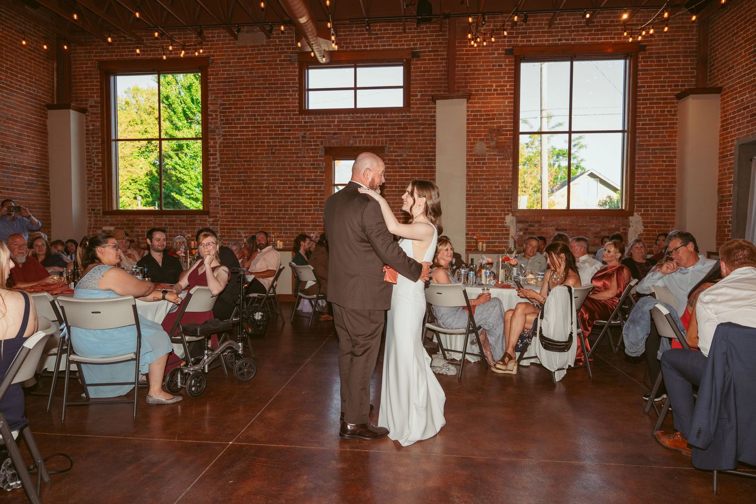 Dancers twirling on wooden floor in rustic brick venue with string lights and tall windows.