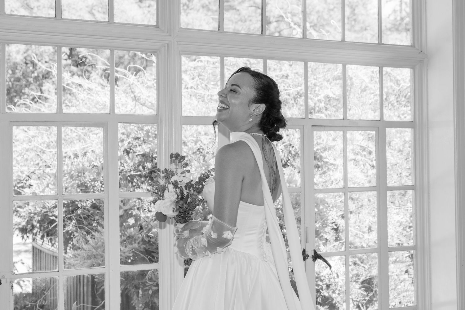 A bride in a classic white wedding dress poses by a sunlit window holding a floral bouquet in black and white.