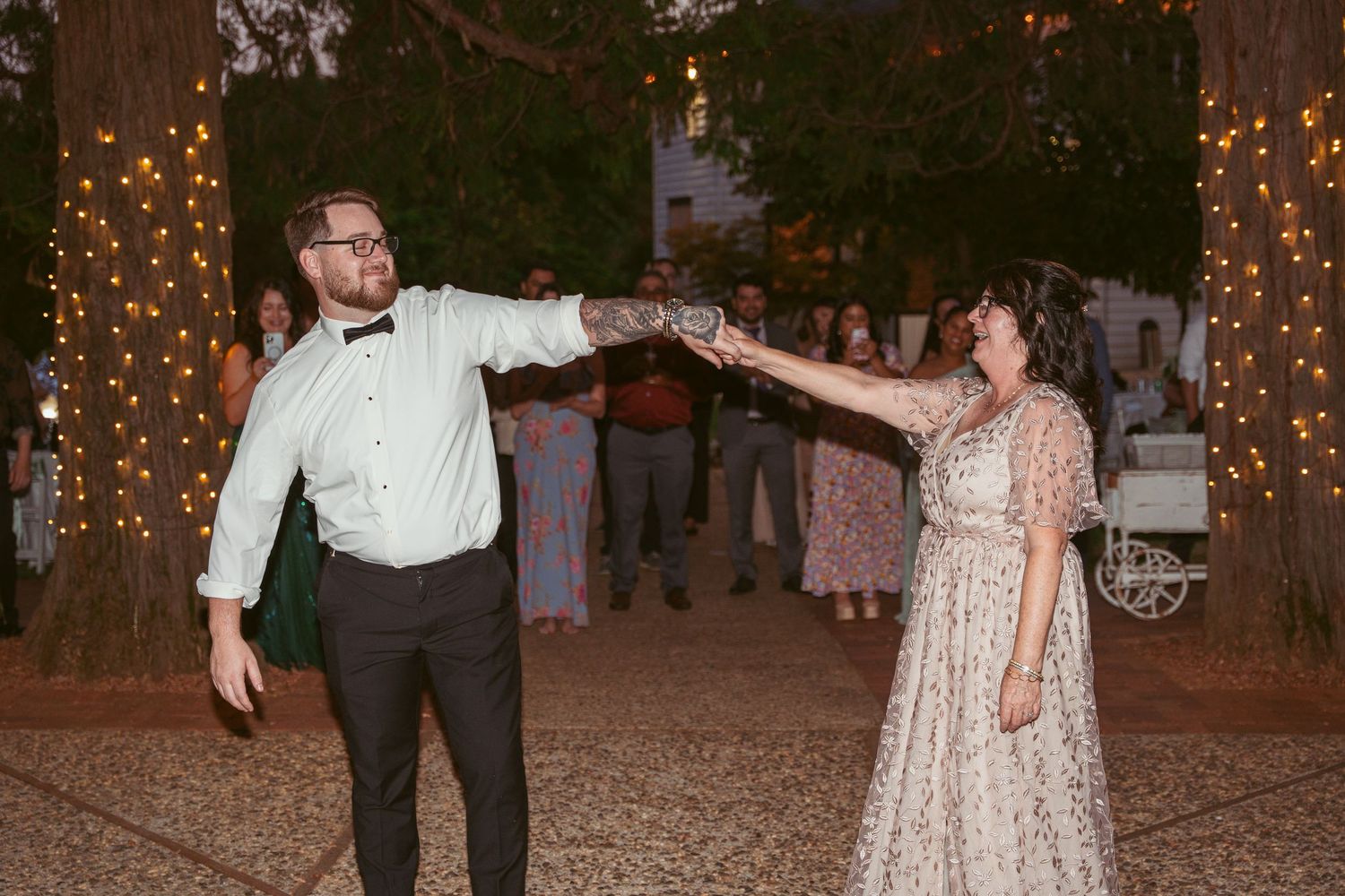 Wedding party dancing and celebrating under string lights at an outdoor evening reception.