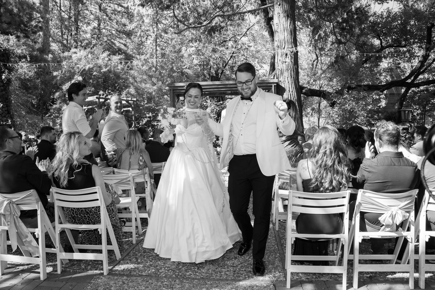 Outdoor wedding ceremony with white chairs arranged in rows under tall trees.