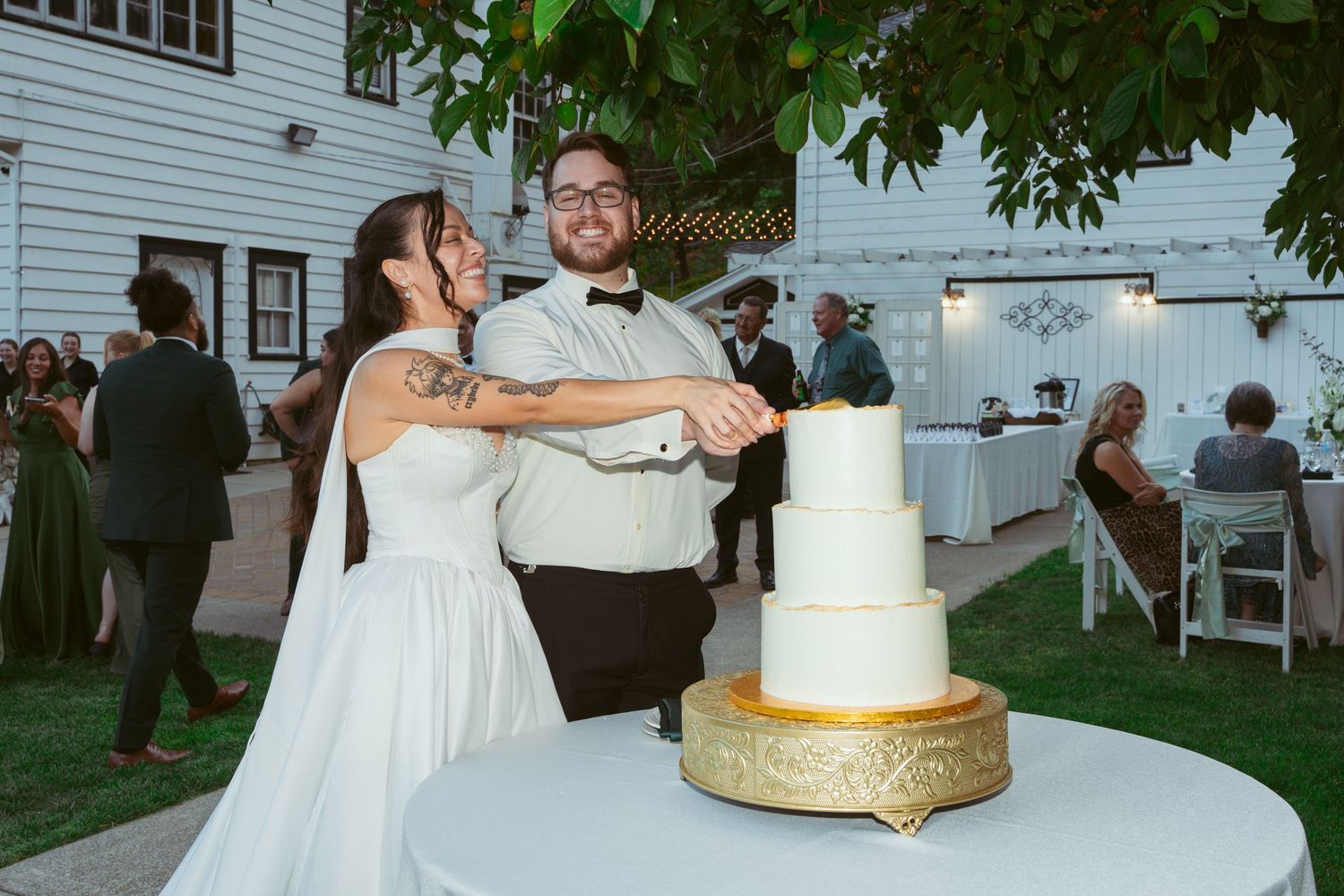 Reception scene with elegant white wedding cake on gold stand as couple celebrates outdoors.