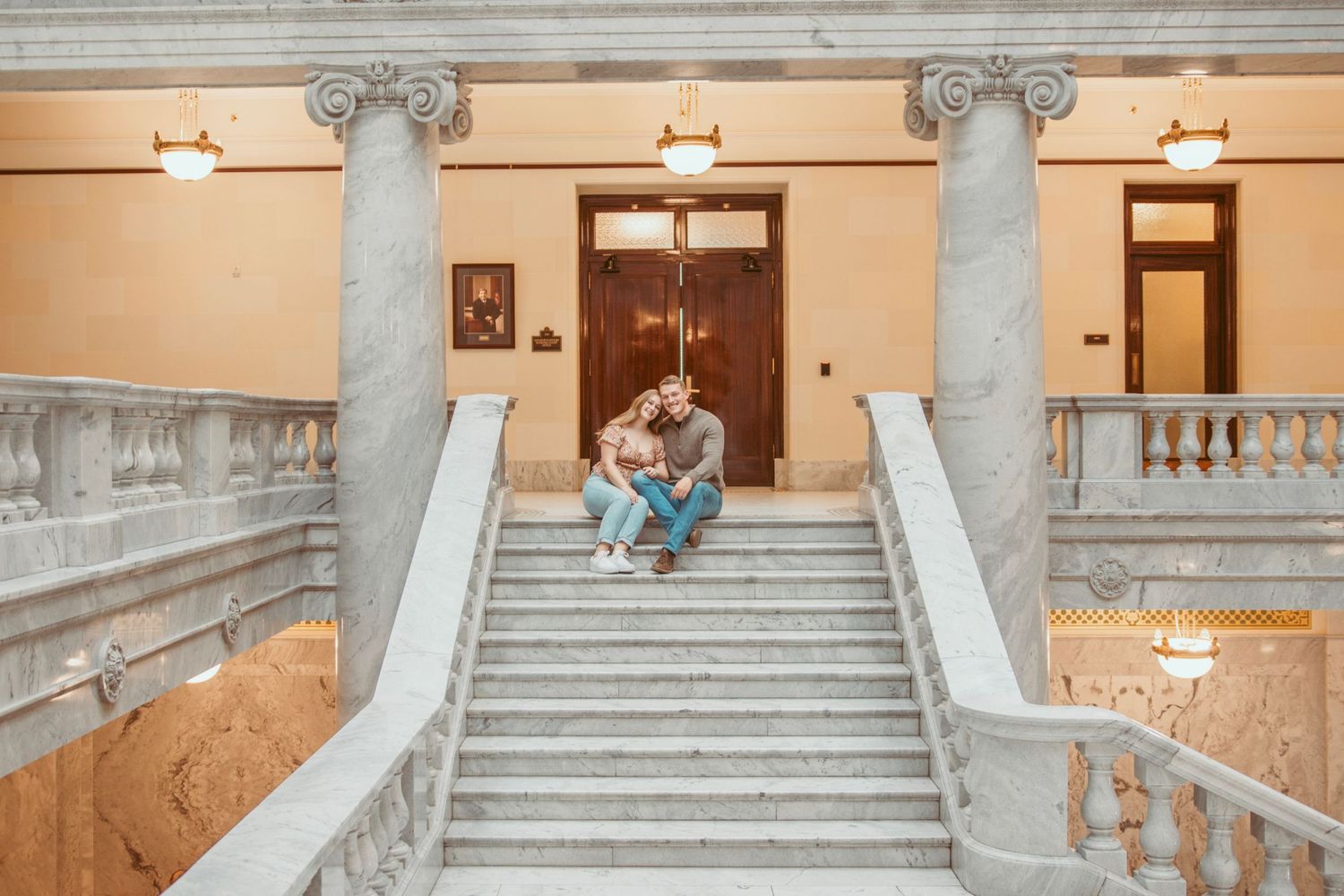 A romantic couple poses on elegant marble stairs in a historic building with ornate architectural details.