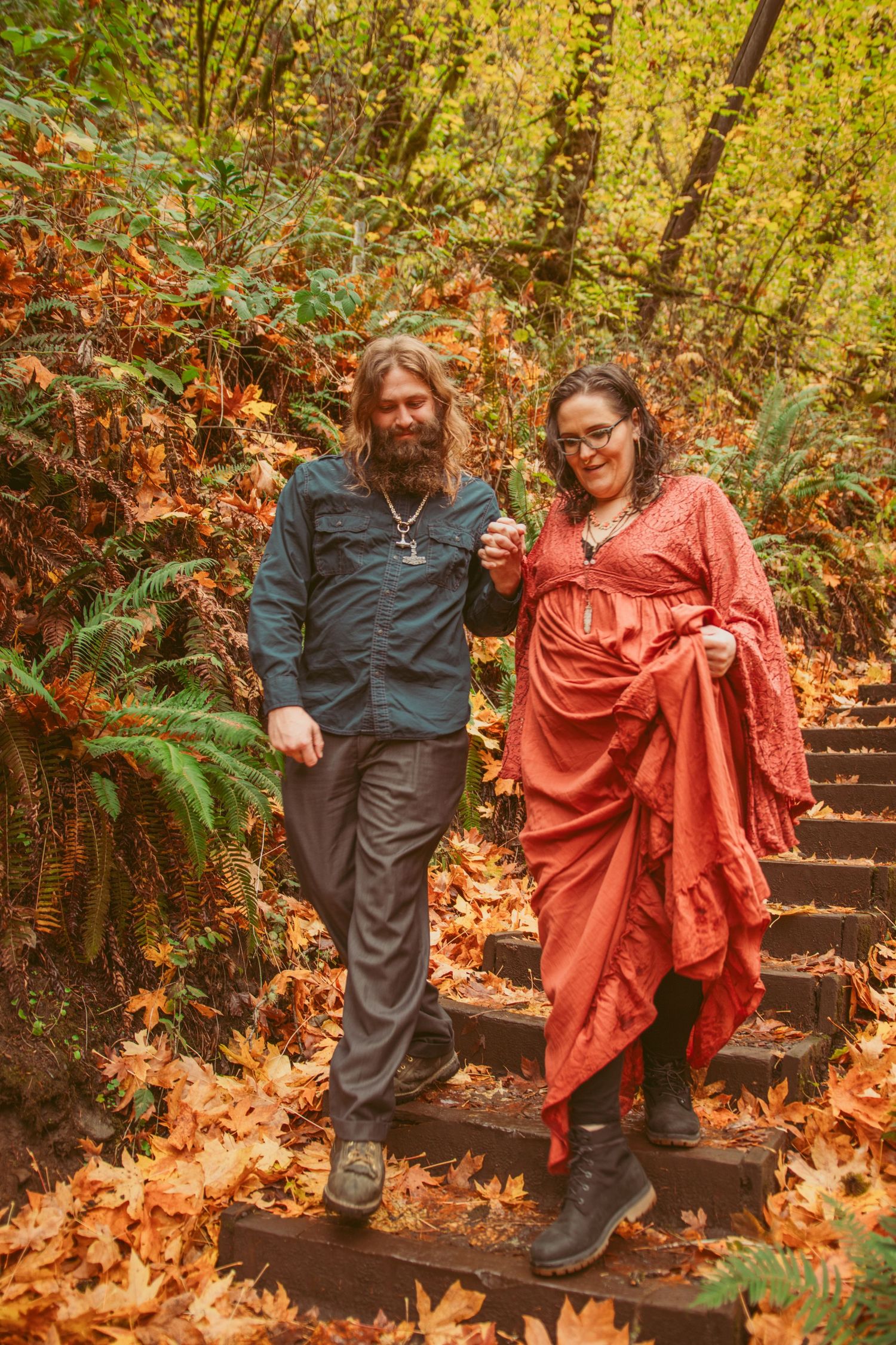 A couple walks down wooden steps covered in autumn leaves while holding each other.