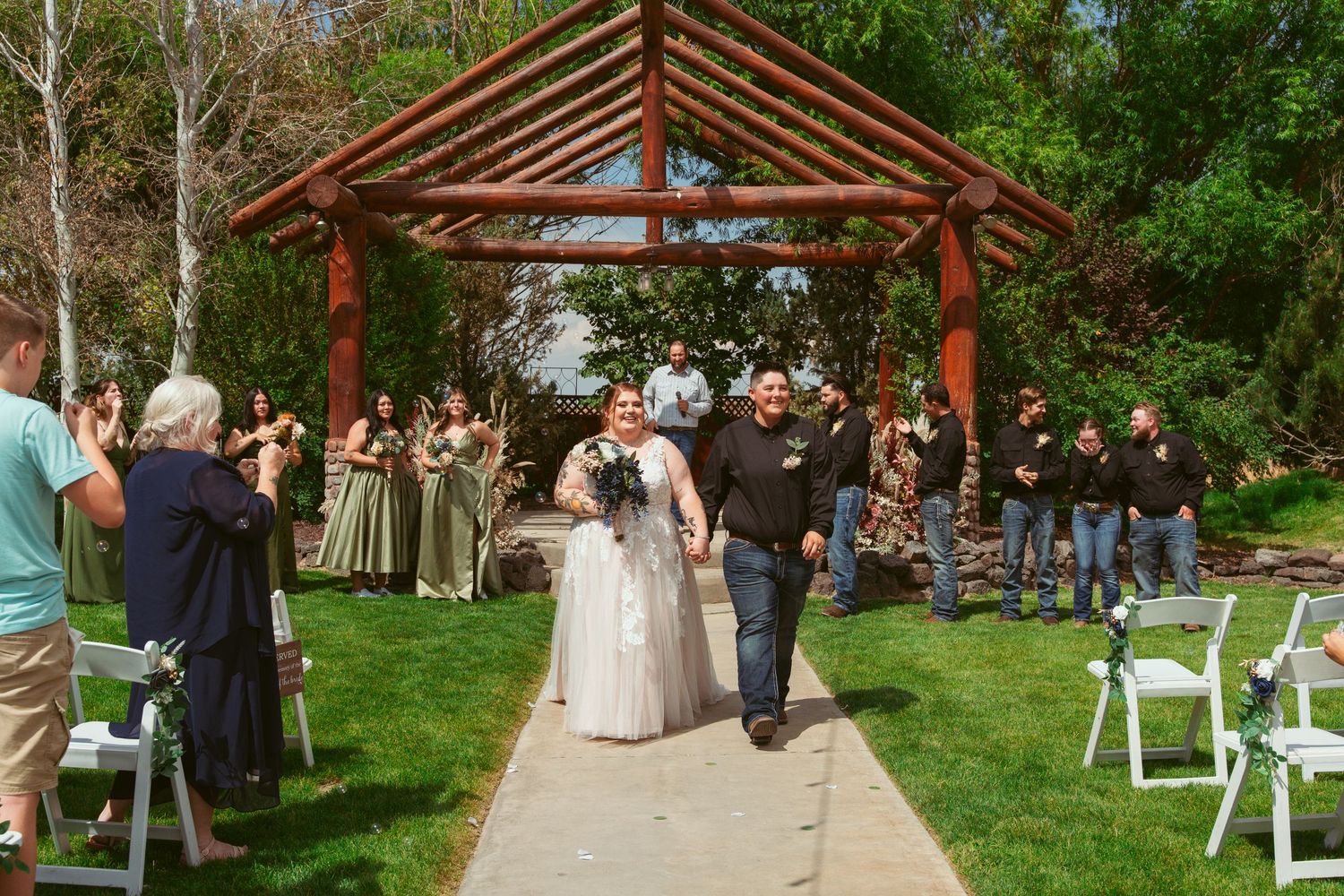 Wedding guests seated in white chairs face a wooden pergola structure during an outdoor ceremony.