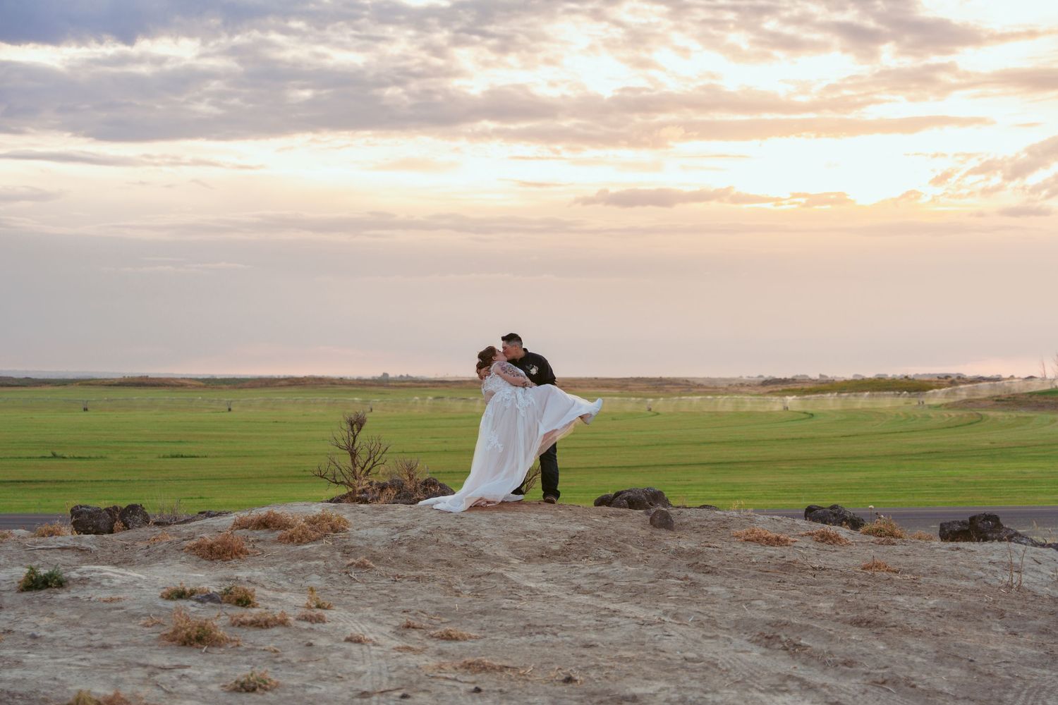 A couple in wedding attire embraces on a hilltop overlooking green fields during a dramatic sunset.