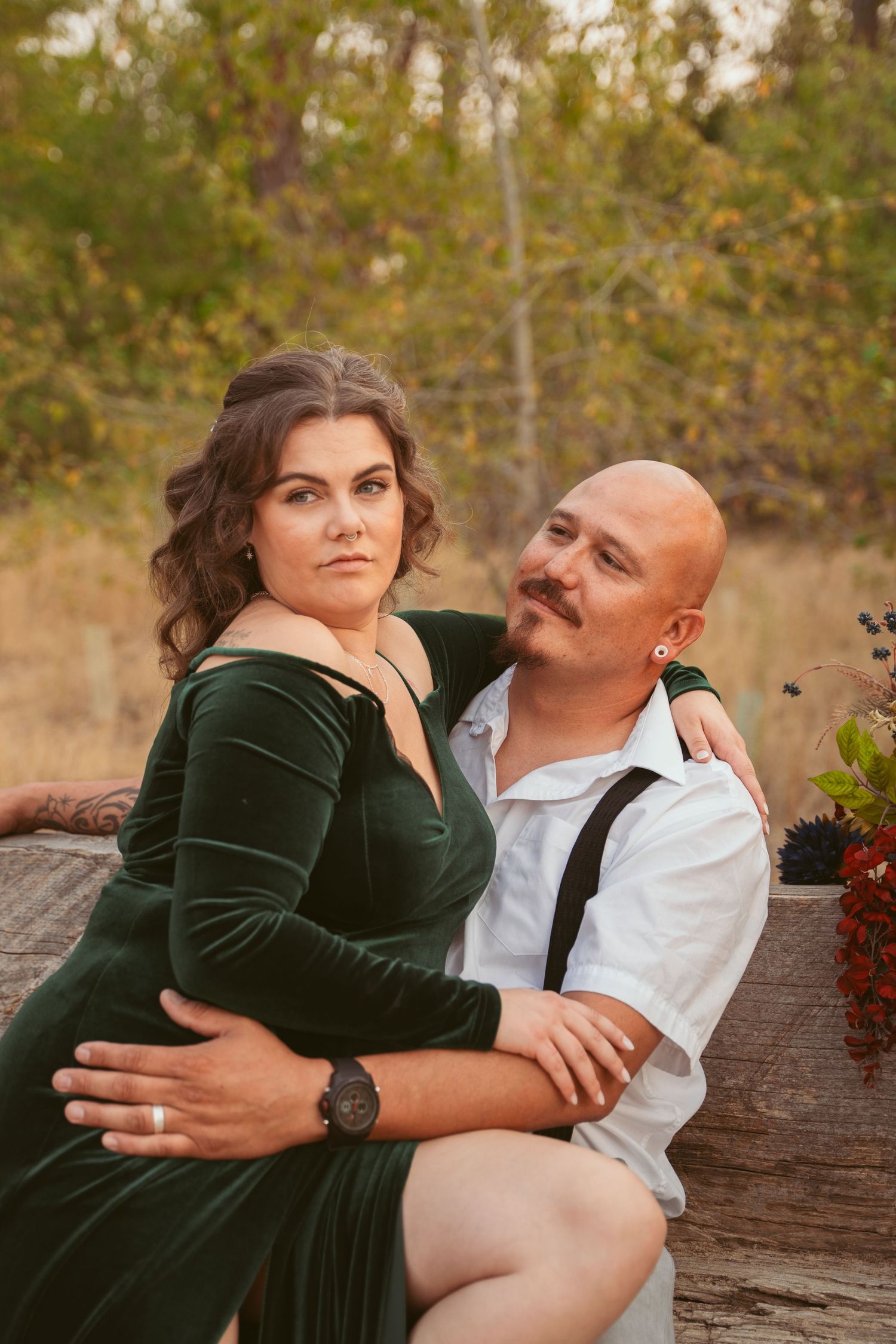 Couple in green velvet dress and white shirt embrace while sitting on a wooden log outdoors.