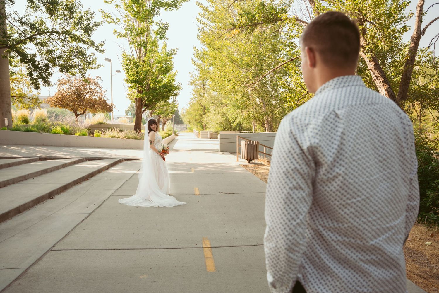 A wedding couple walking together on a paved path surrounded by green trees and sunlight.
