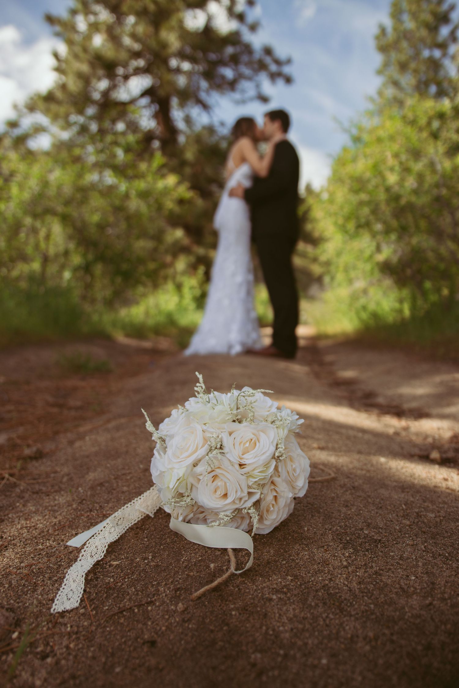 White rose bridal bouquet on dirt path with couple in soft focus background.