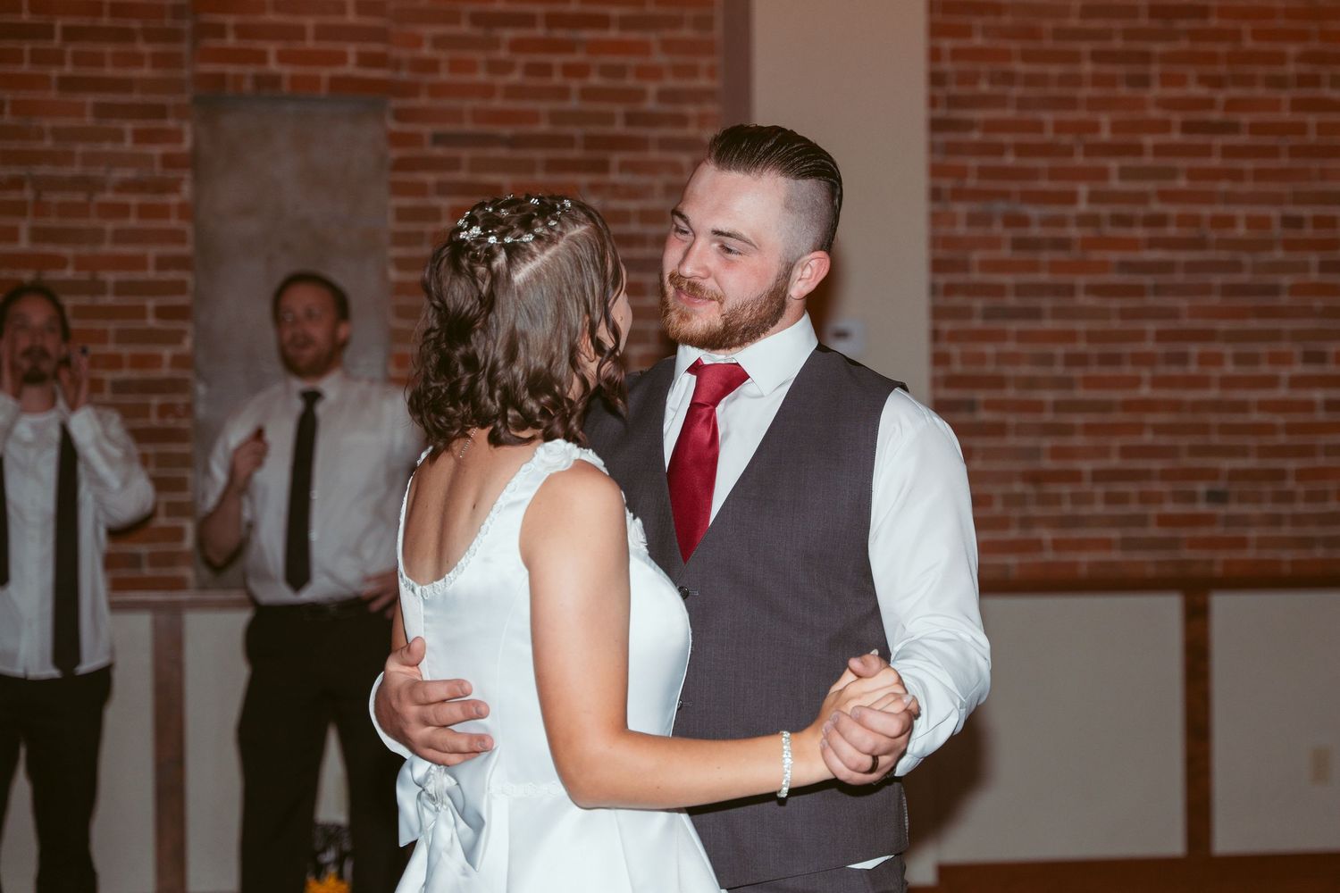 Couple dancing together in a rustic brick-walled venue with string lights overhead.