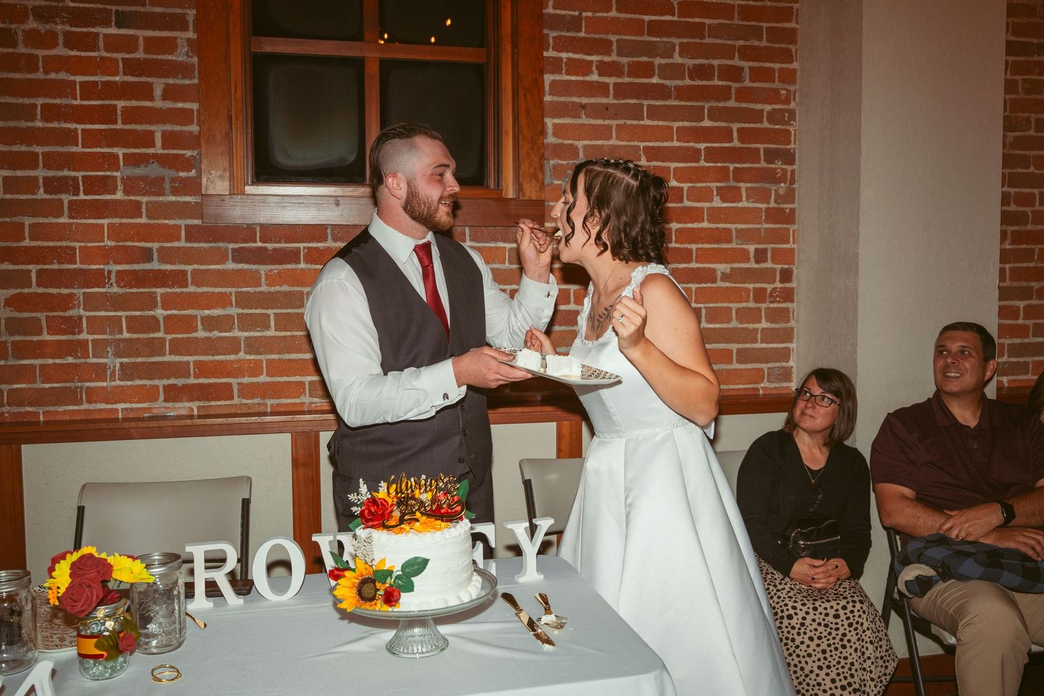 Newlyweds share a fun cake-feeding moment during their reception in a rustic brick venue.