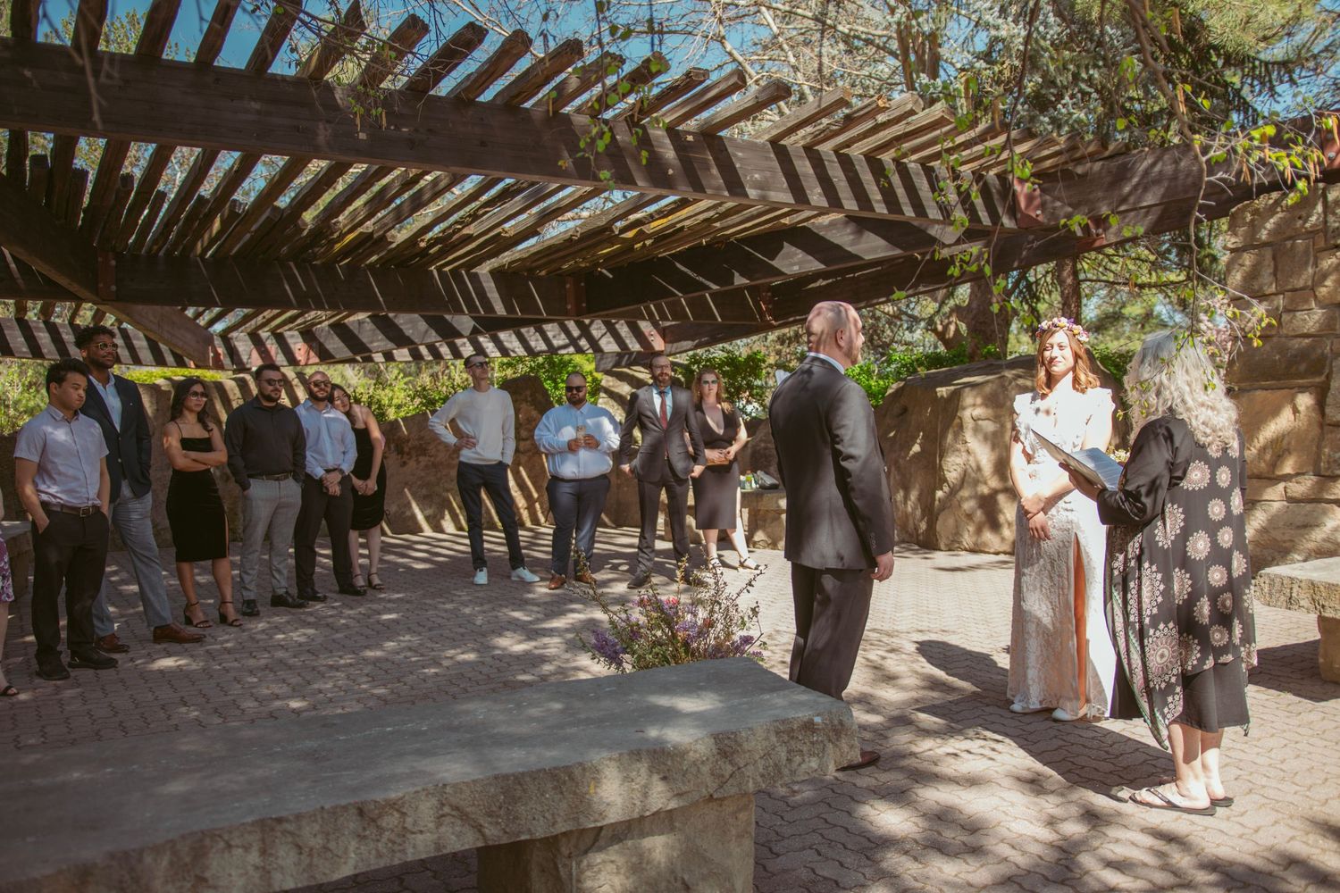 A wedding ceremony takes place under a wooden pergola with guests gathered around.