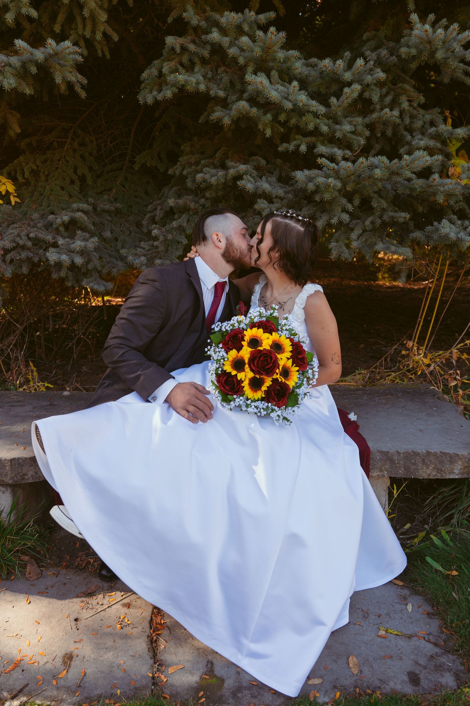 Newlyweds share romantic moment on stone bench with sunflower bouquet and flowing white dress.