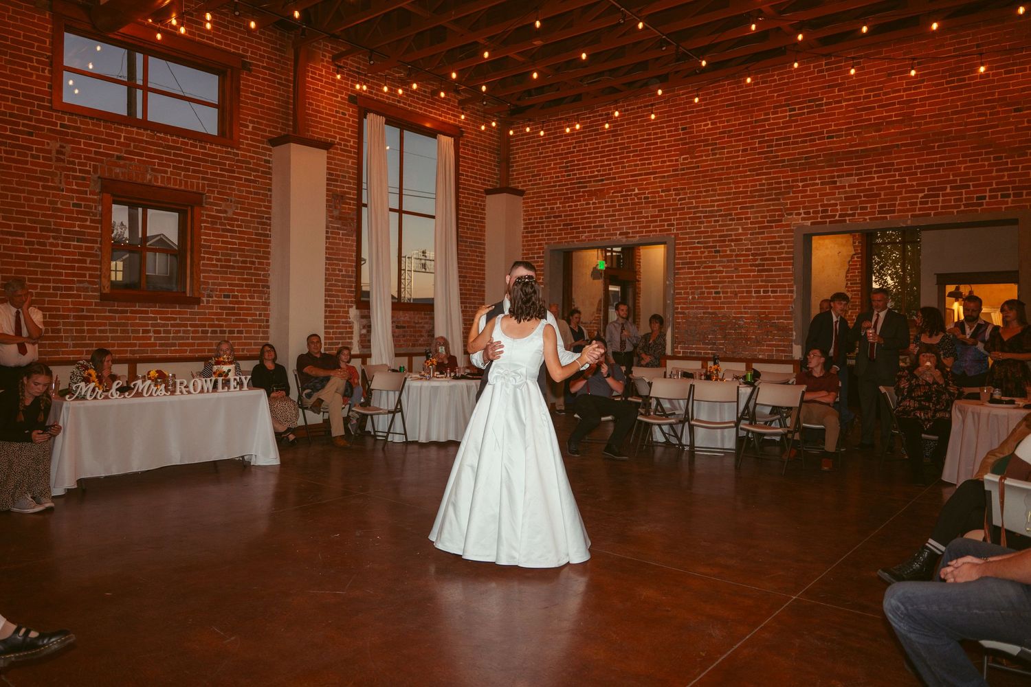 Wedding couple dances beneath twinkling lights in a rustic brick reception venue.