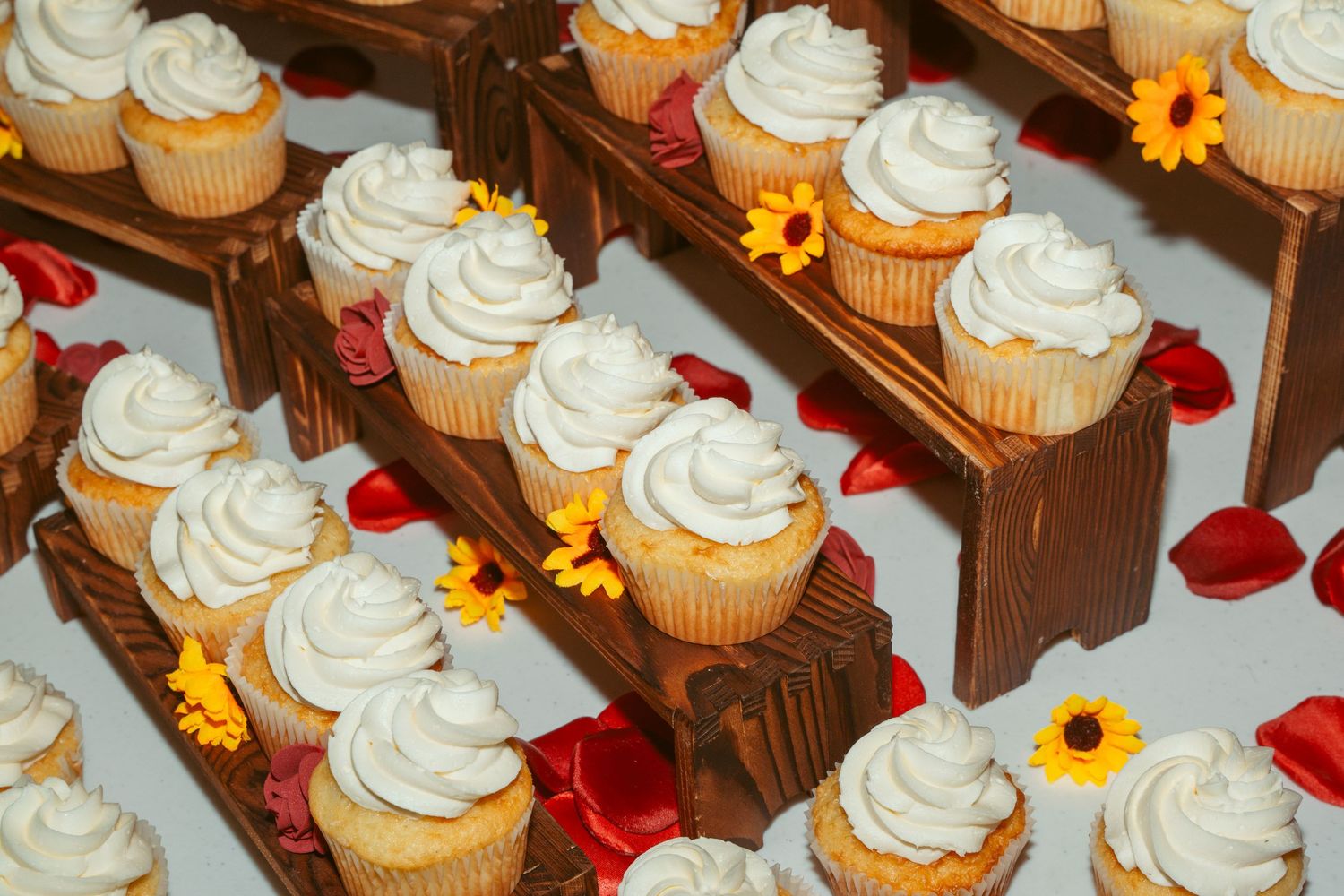Rows of vanilla cupcakes with white frosting swirls decorated with red and yellow flower petals.