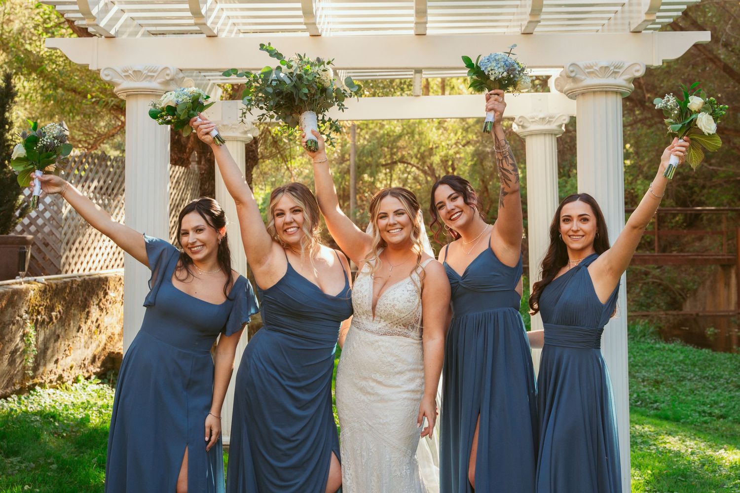 Wedding party poses together under a white pergola surrounded by green grass and trees.