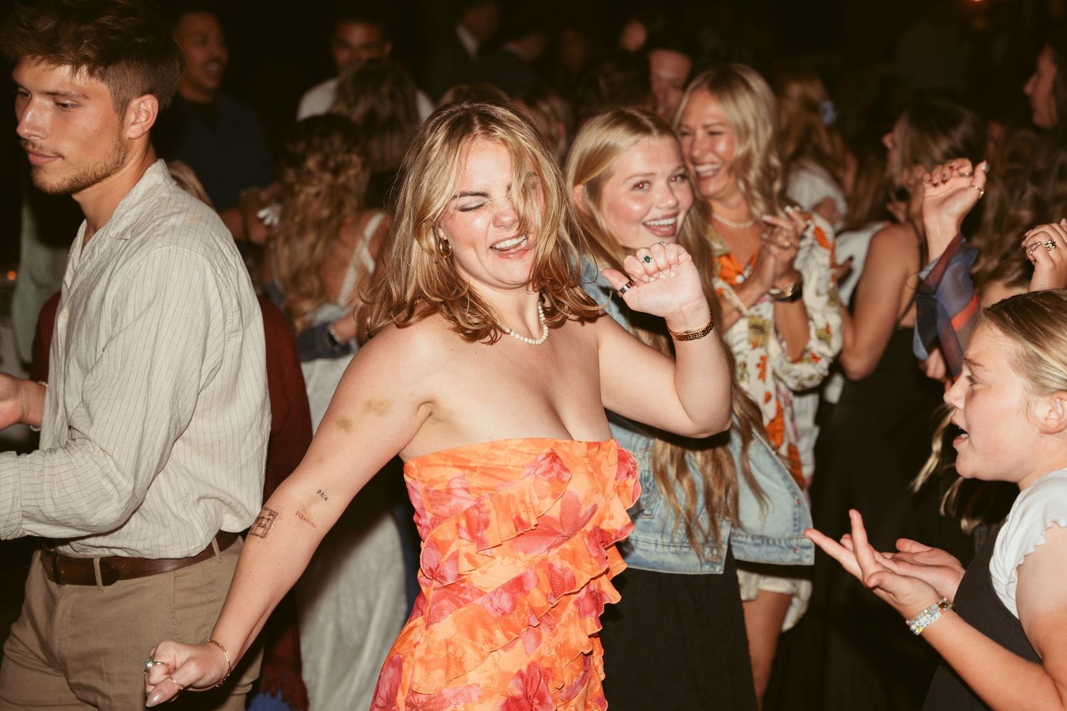 Guests dancing and celebrating at a lively party with someone in an orange floral dress in the foreground.