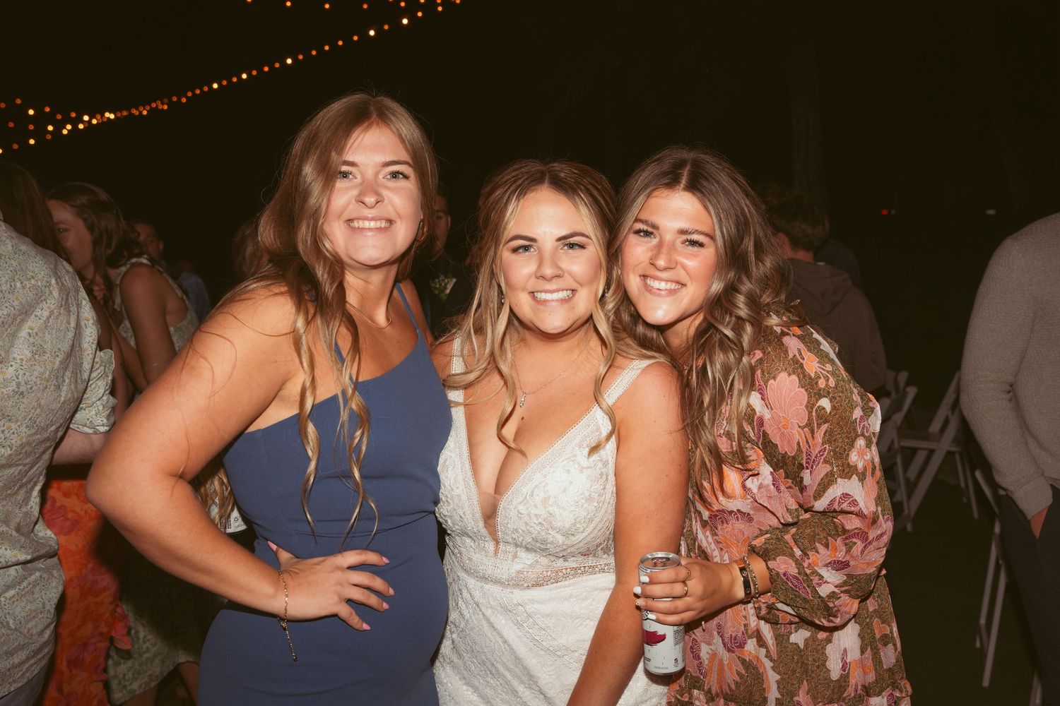 A group of friends in formal attire pose together at a nighttime celebration event with string lights in the background.