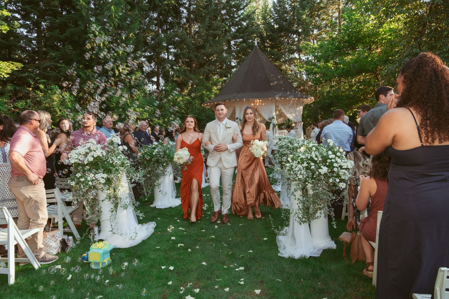 Wedding party stands at garden gazebo surrounded by white floral arrangements and seated guests.