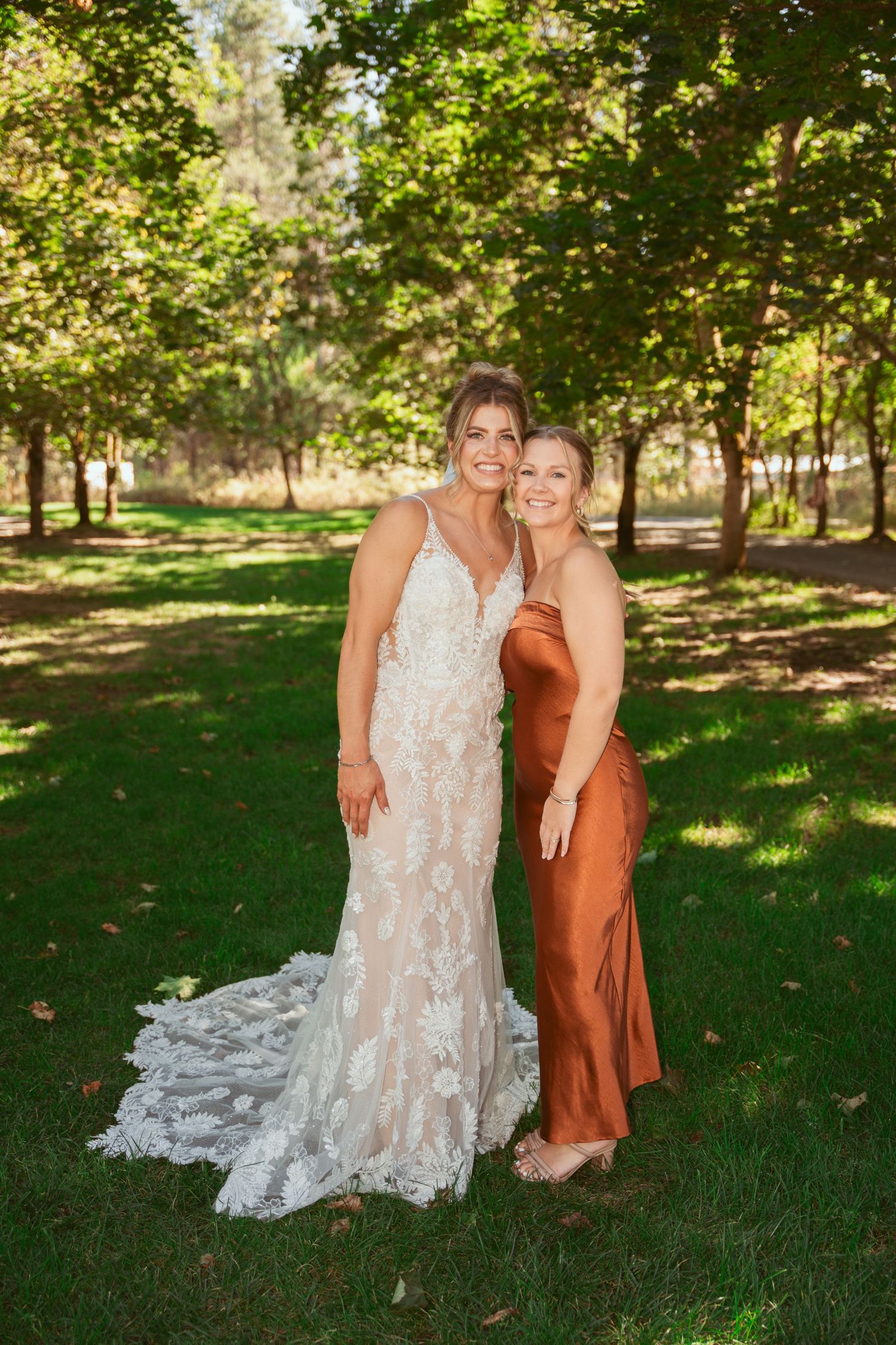 Bridal party members in rust colored dresses gather for photos on green lawn under trees.