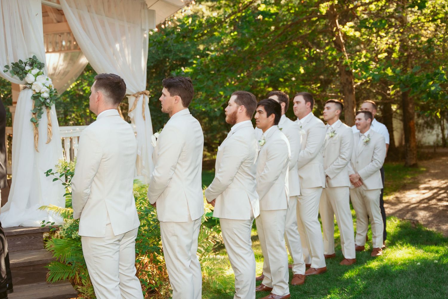 Wedding party members in white attire lined up outdoors near a gazebo.
