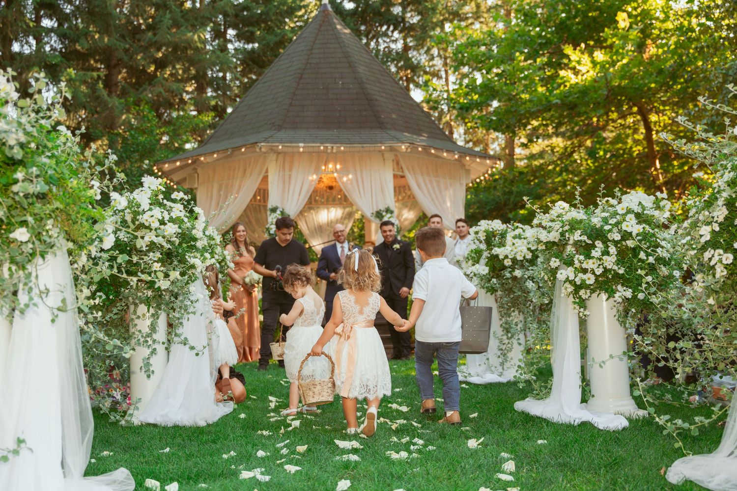 A string of outdoor wedding tents with white fabric draped between trees creates an enchanting garden ceremony setting.