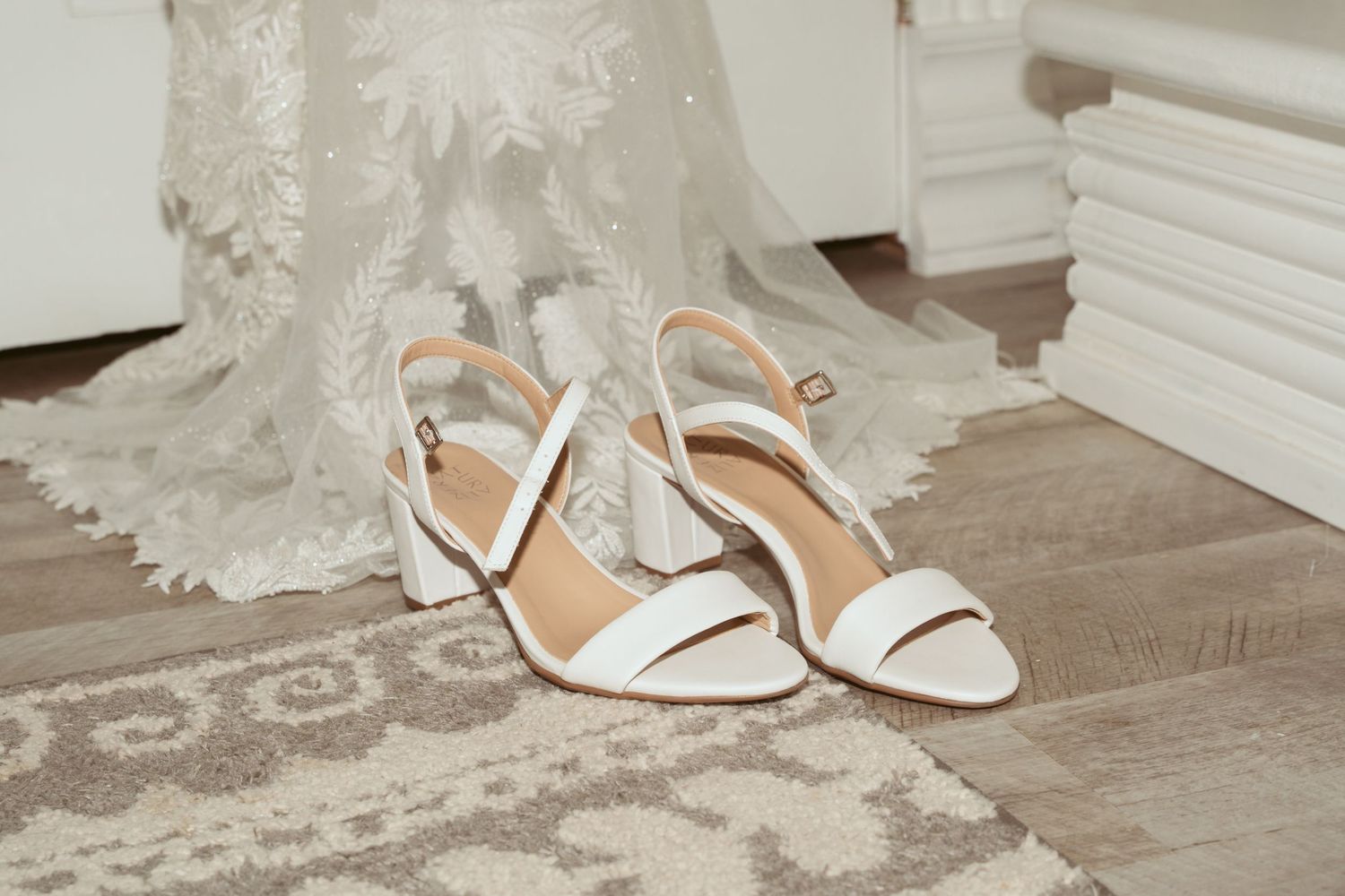 Elegant white bridal sandals with delicate straps sit beside a lace wedding dress on a wooden floor.