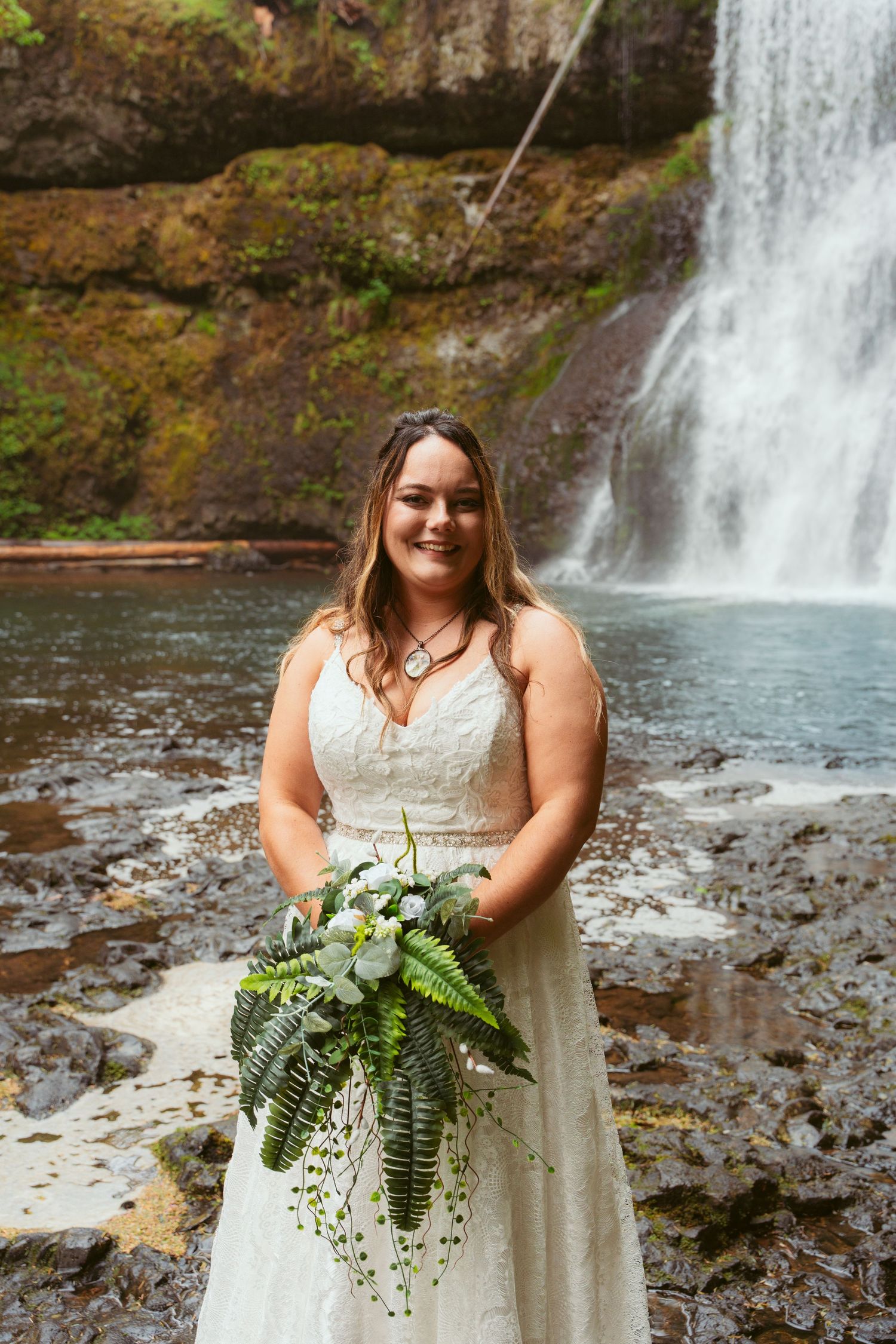 Elegant bridal portrait showcasing a white lace gown and lush greenery bouquet by a waterfall.