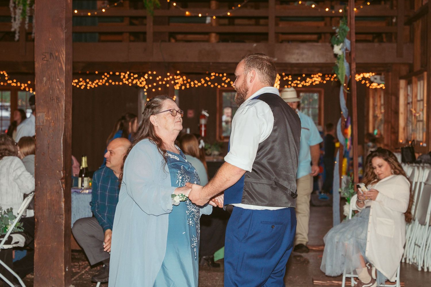 Party guests dance and celebrate in a rustic barn setting with string light decorations.