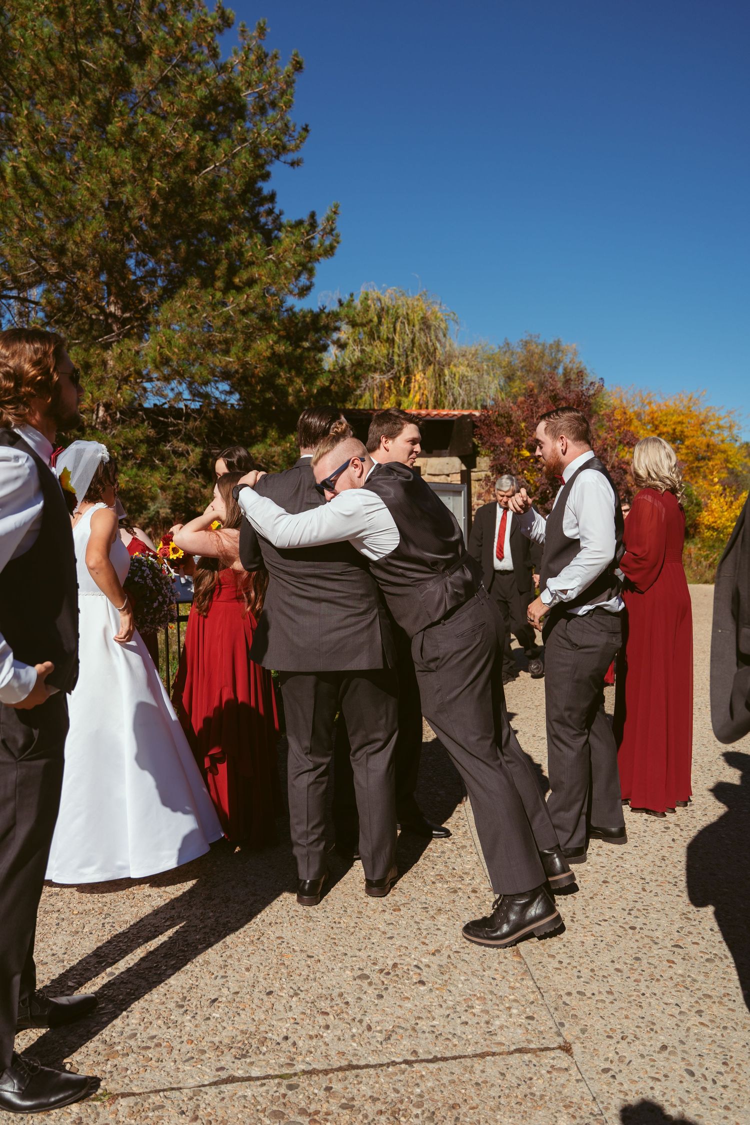 Wedding party lined up for creative photo on gravel path with pine trees in background.