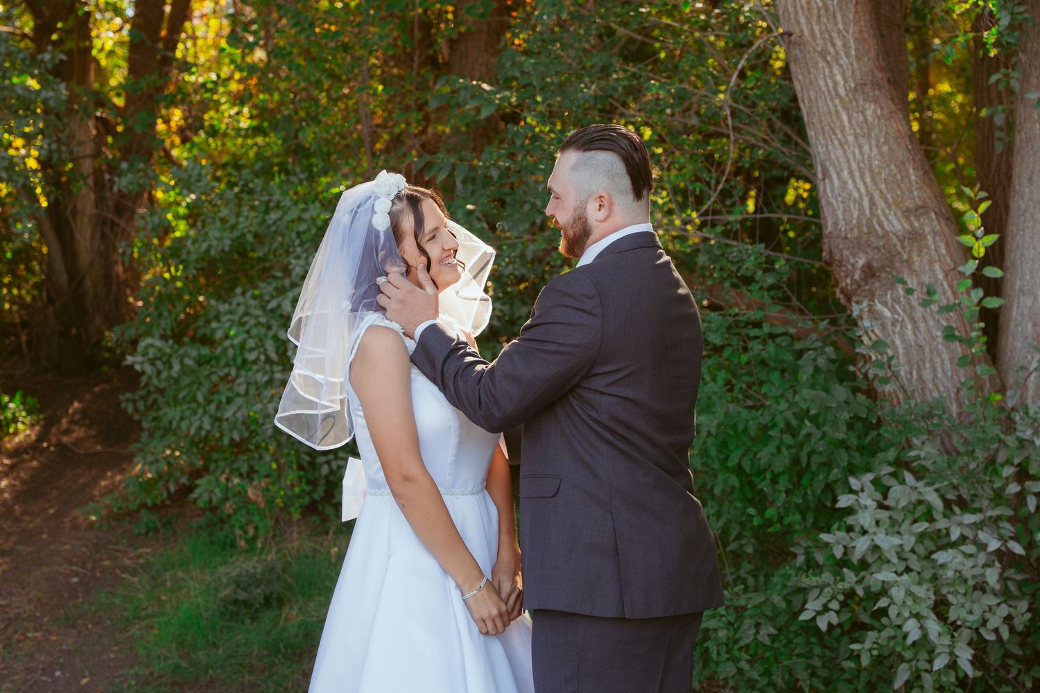 Couple shares an intimate moment during their wedding photoshoot in a sunlit wooded setting.