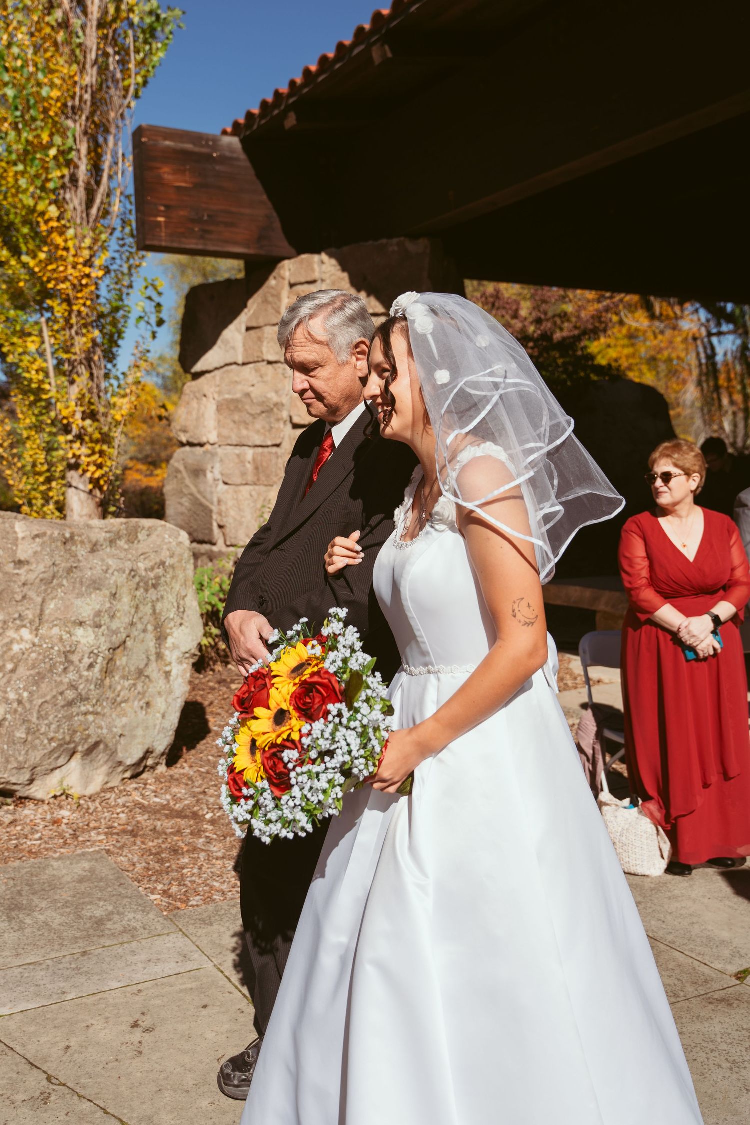 Bride in white dress carries sunflower bouquet during autumn outdoor ceremony.