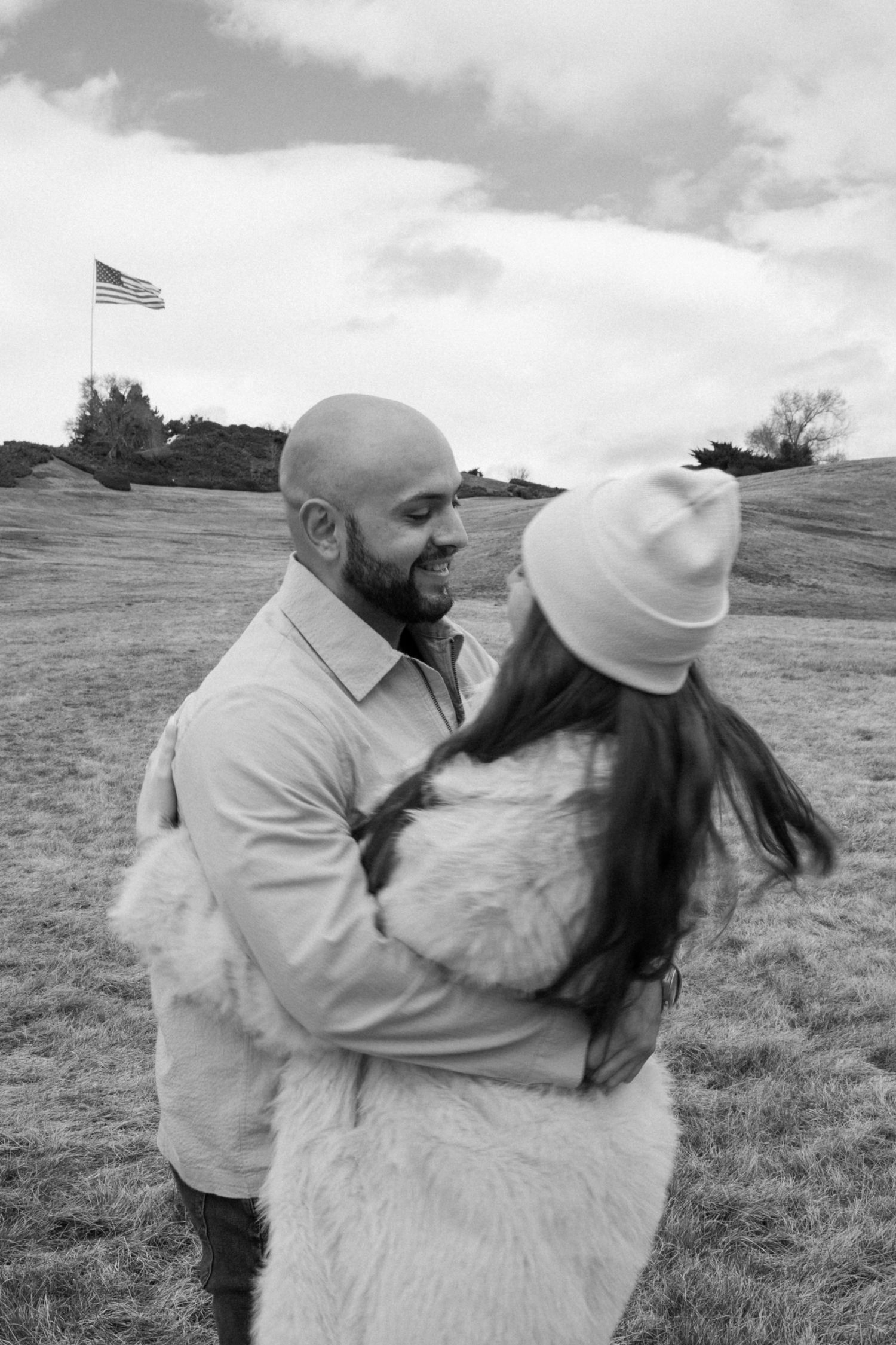 Playful couple dancing and embracing in an outdoor field with American flag in background.
