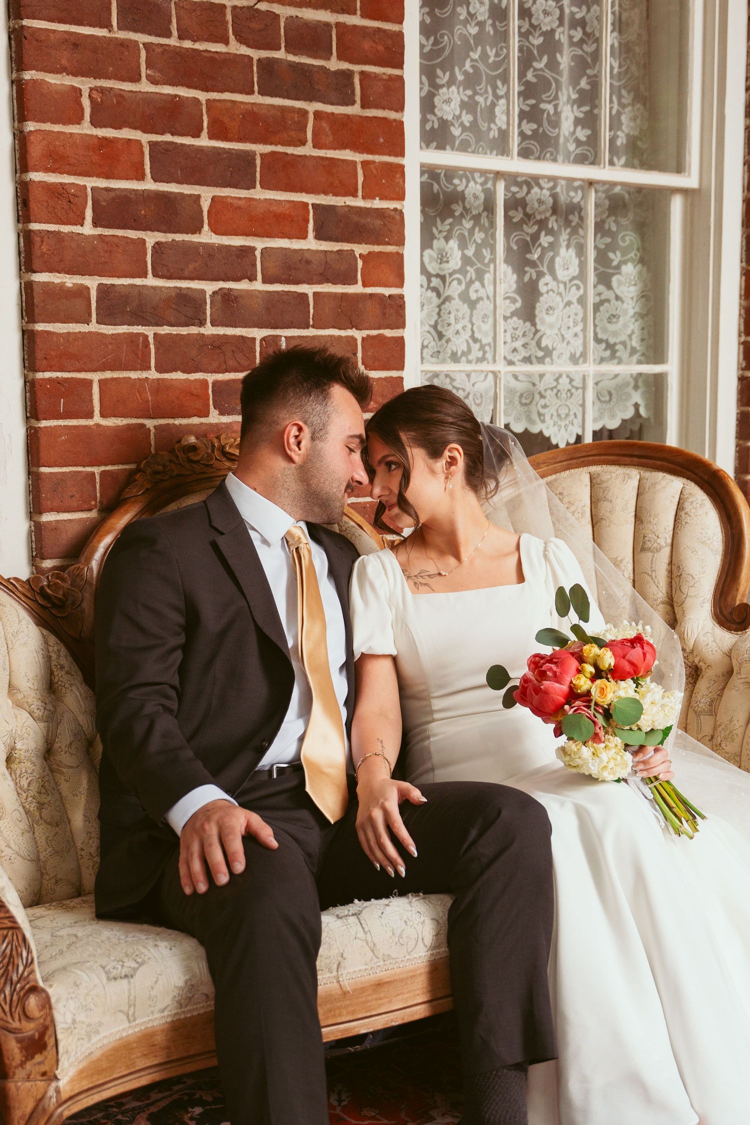 A romantic wedding portrait of a couple sitting on a vintage sofa against a brick wall.