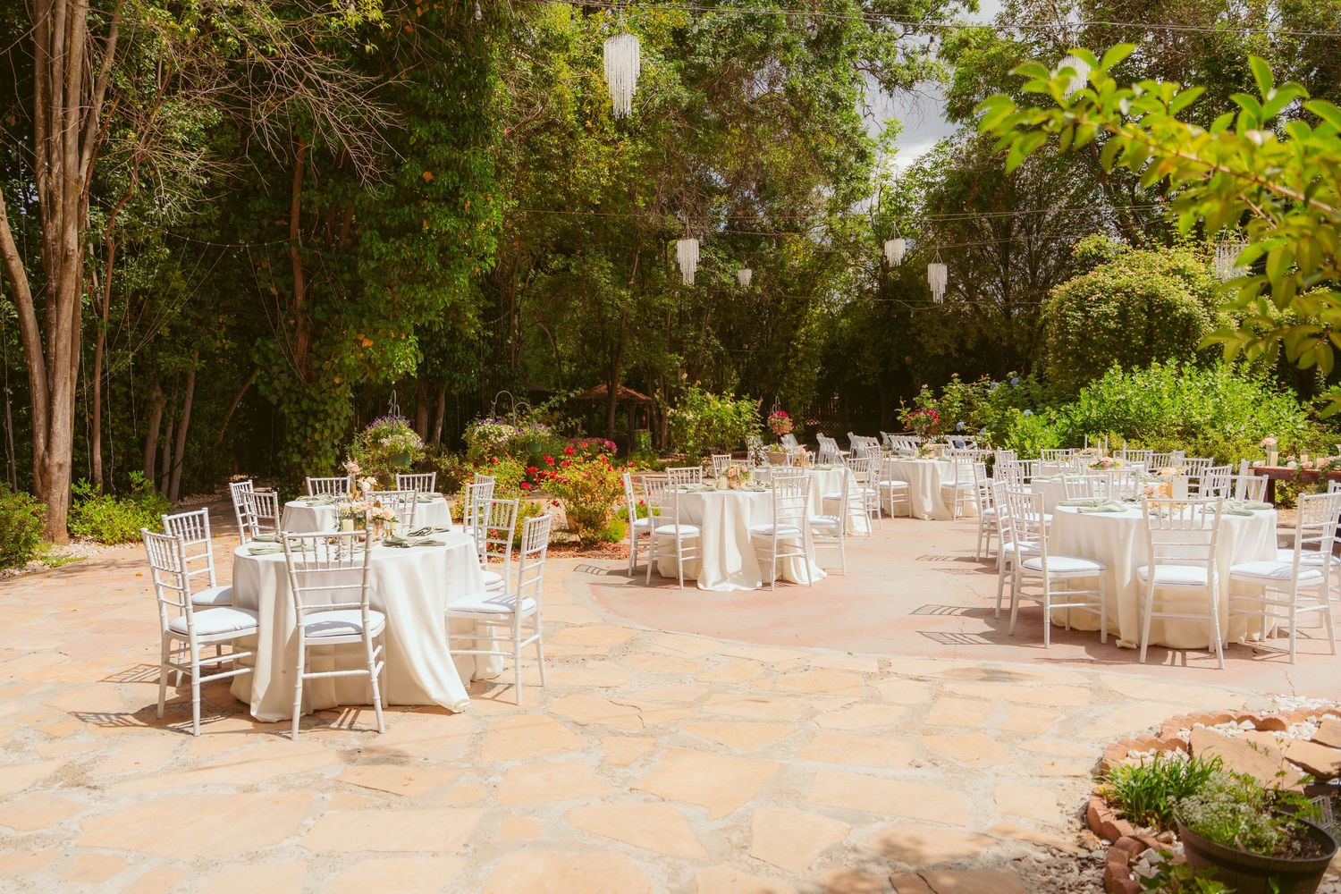 A garden wedding reception area with white chairs and tables arranged under tree canopies.