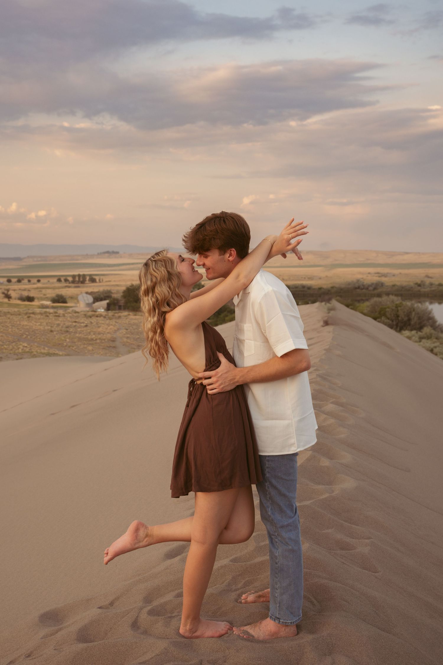 A playful couple embraces while standing on desert sand dunes at sunset.