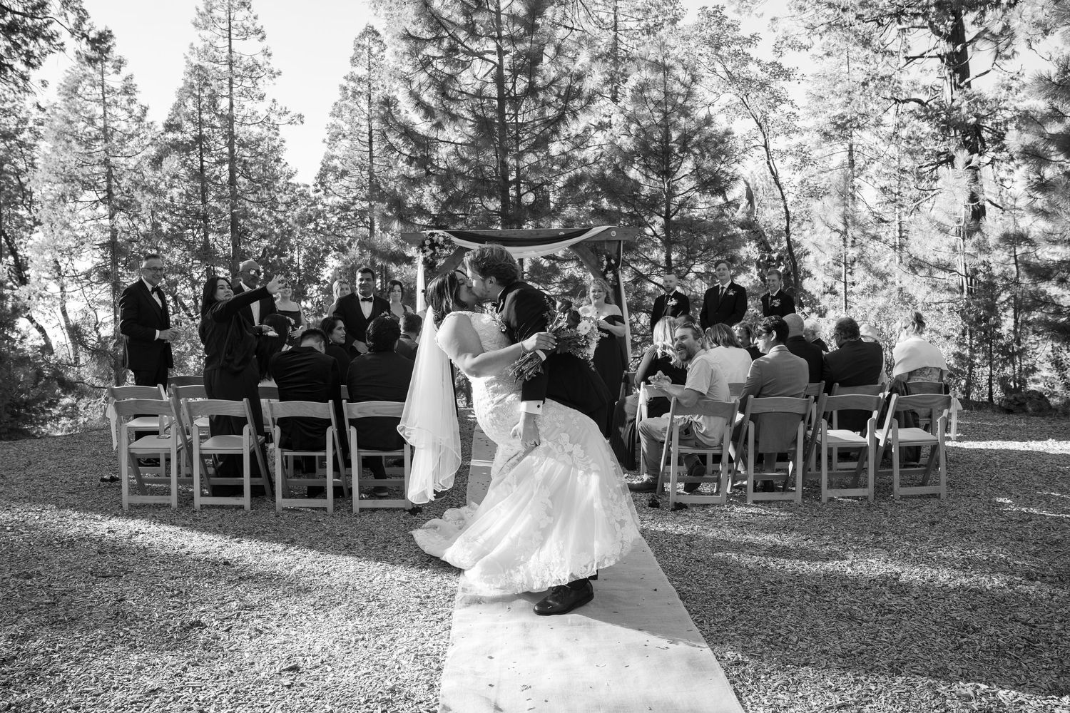 Wedding ceremony taking place outdoors in a wooded setting with wooden chairs and natural decor.
