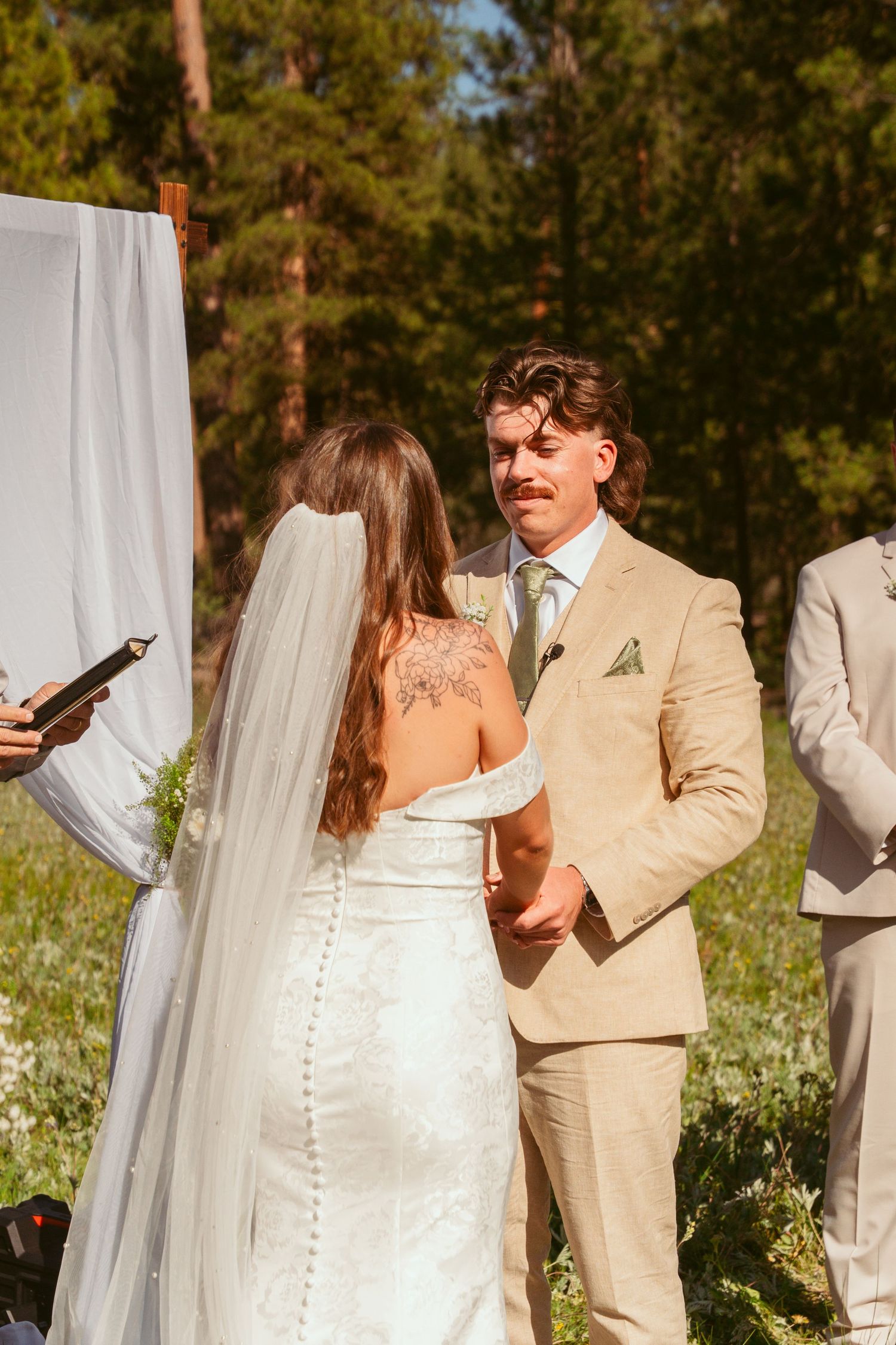 Outdoor wedding ceremony in a forest setting with white fabric draping and light tan suits.