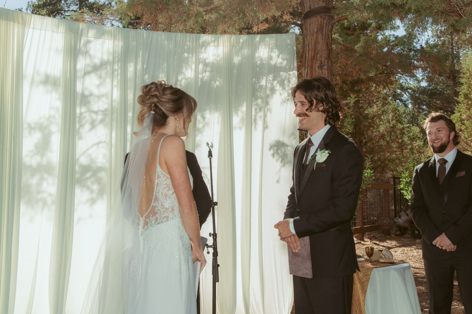 Wedding ceremony in progress under white drapes and towering pine trees.