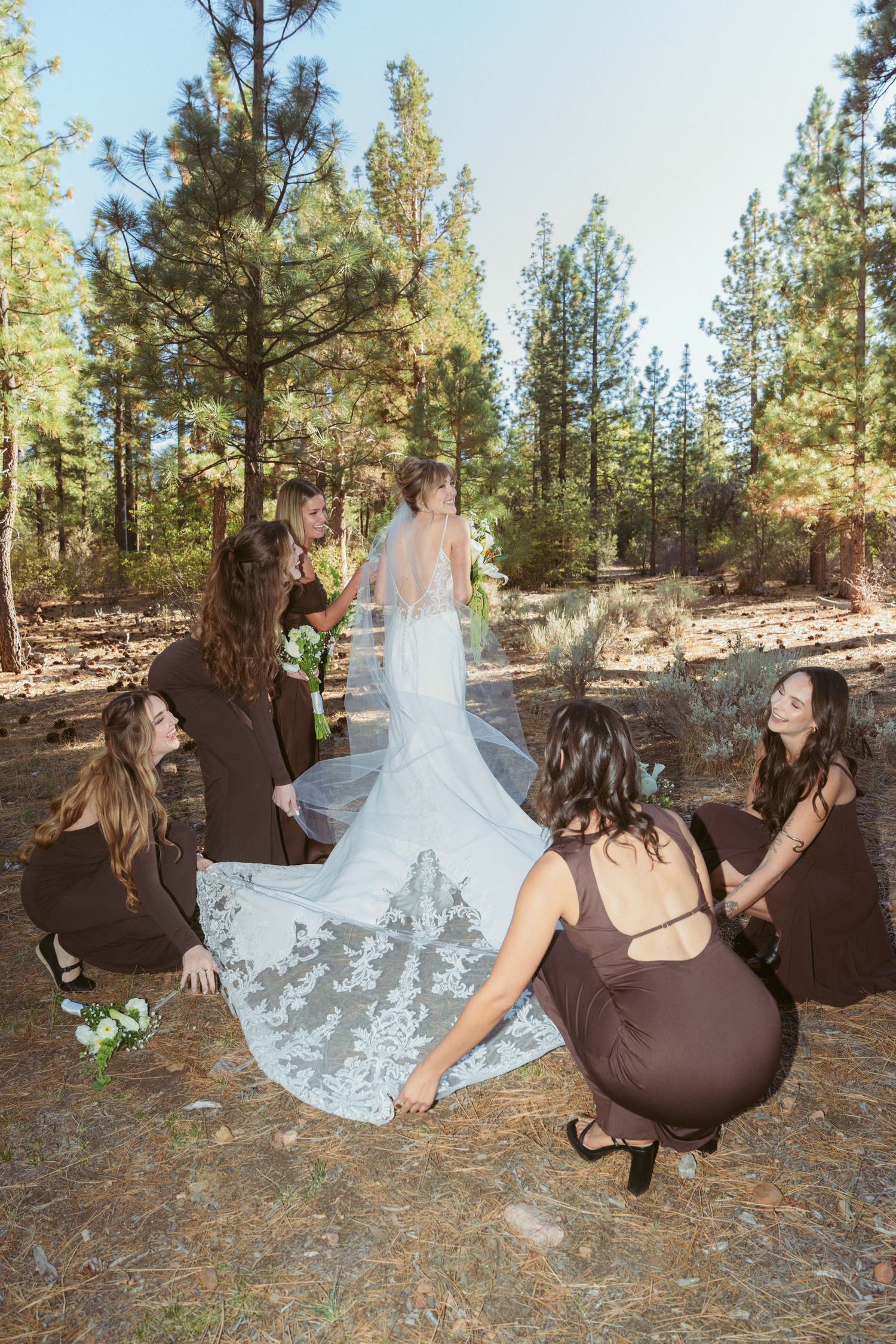 Wedding party members in brown dresses create fun falling poses on forest ground.