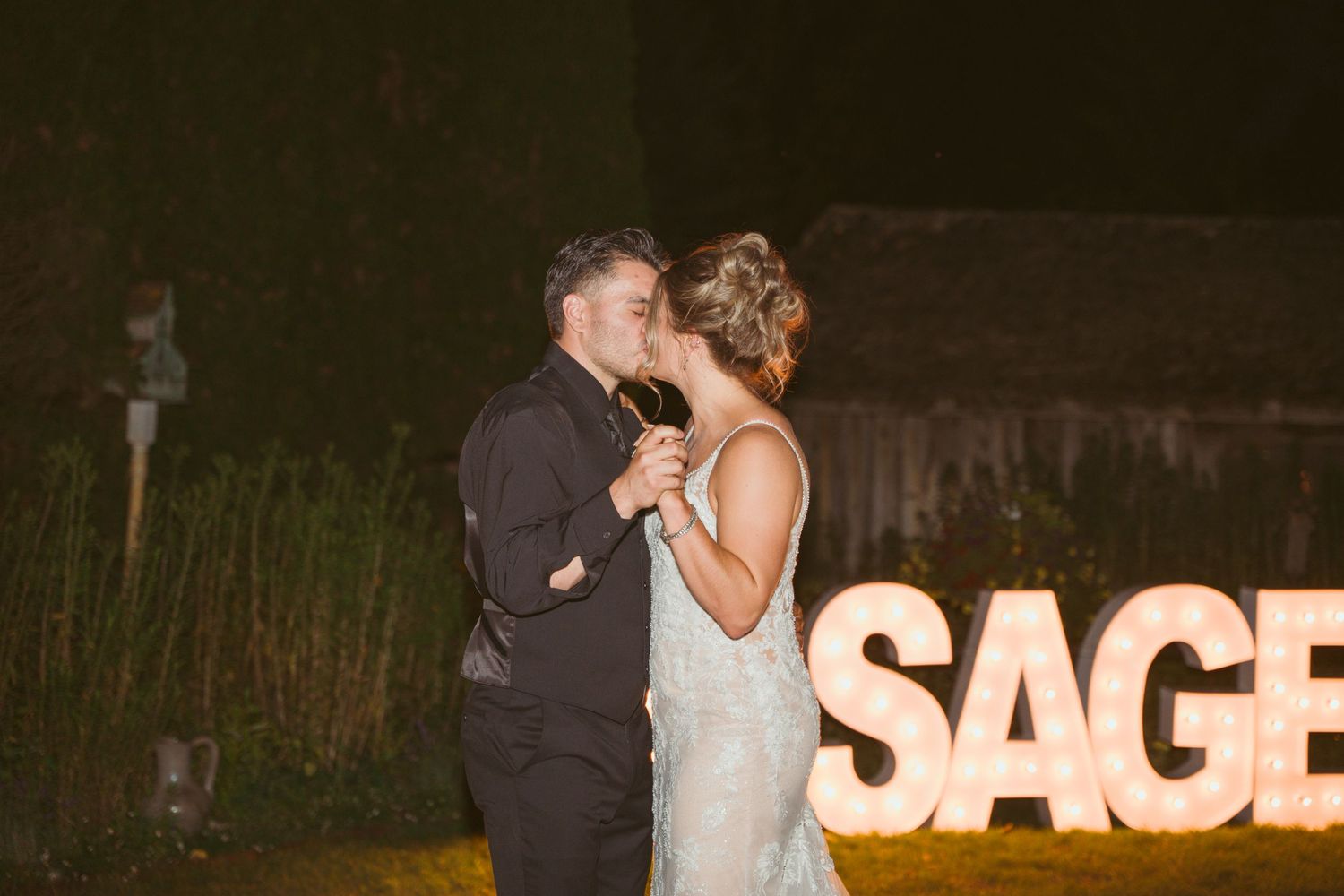 Couple dancing at night in front of illuminated SAGE letters during wedding reception.